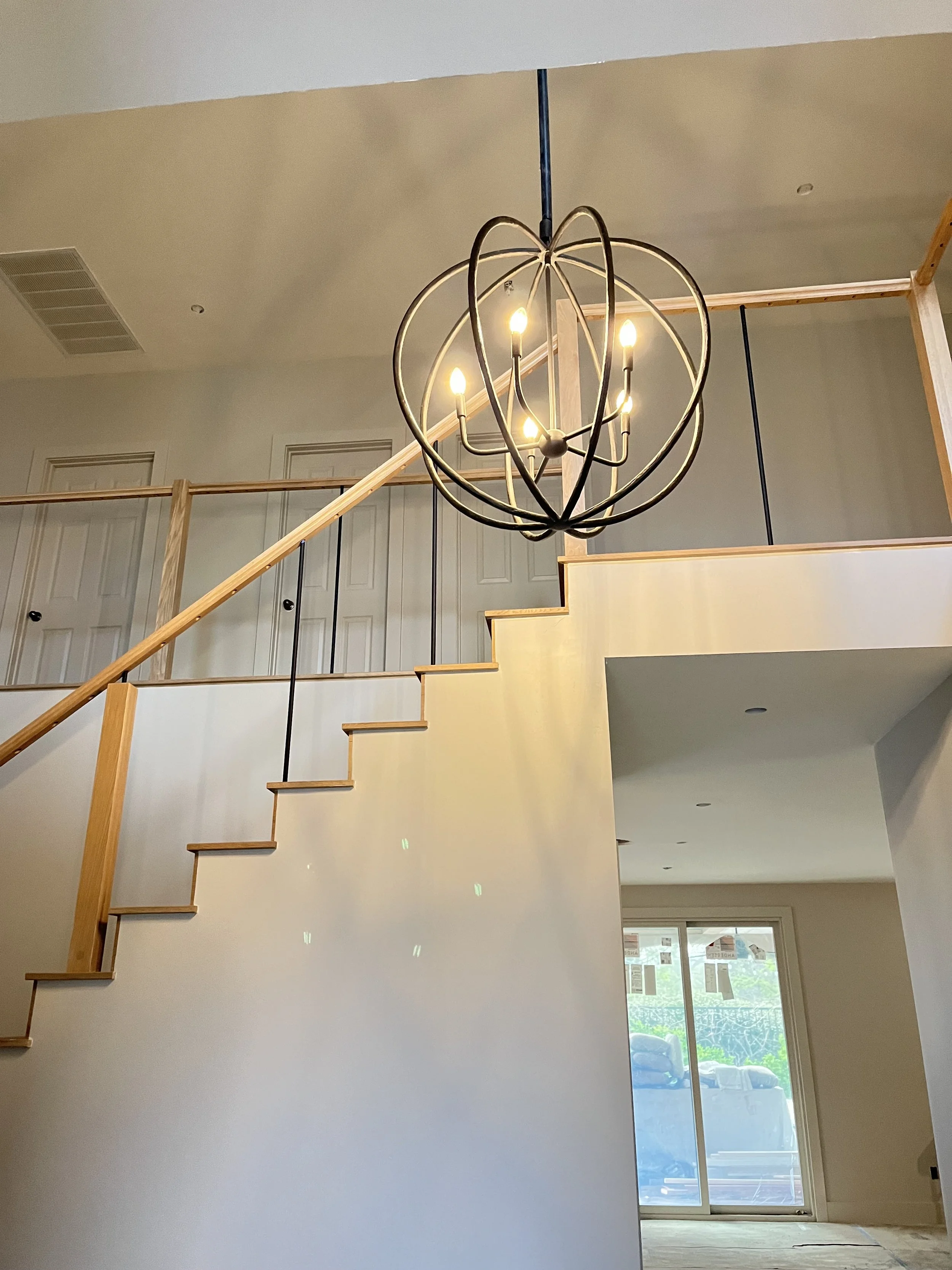 Interior of a modern home under construction with a staircase, wooden railing, a large round chandelier with five candles, and a sliding glass door leading outside.