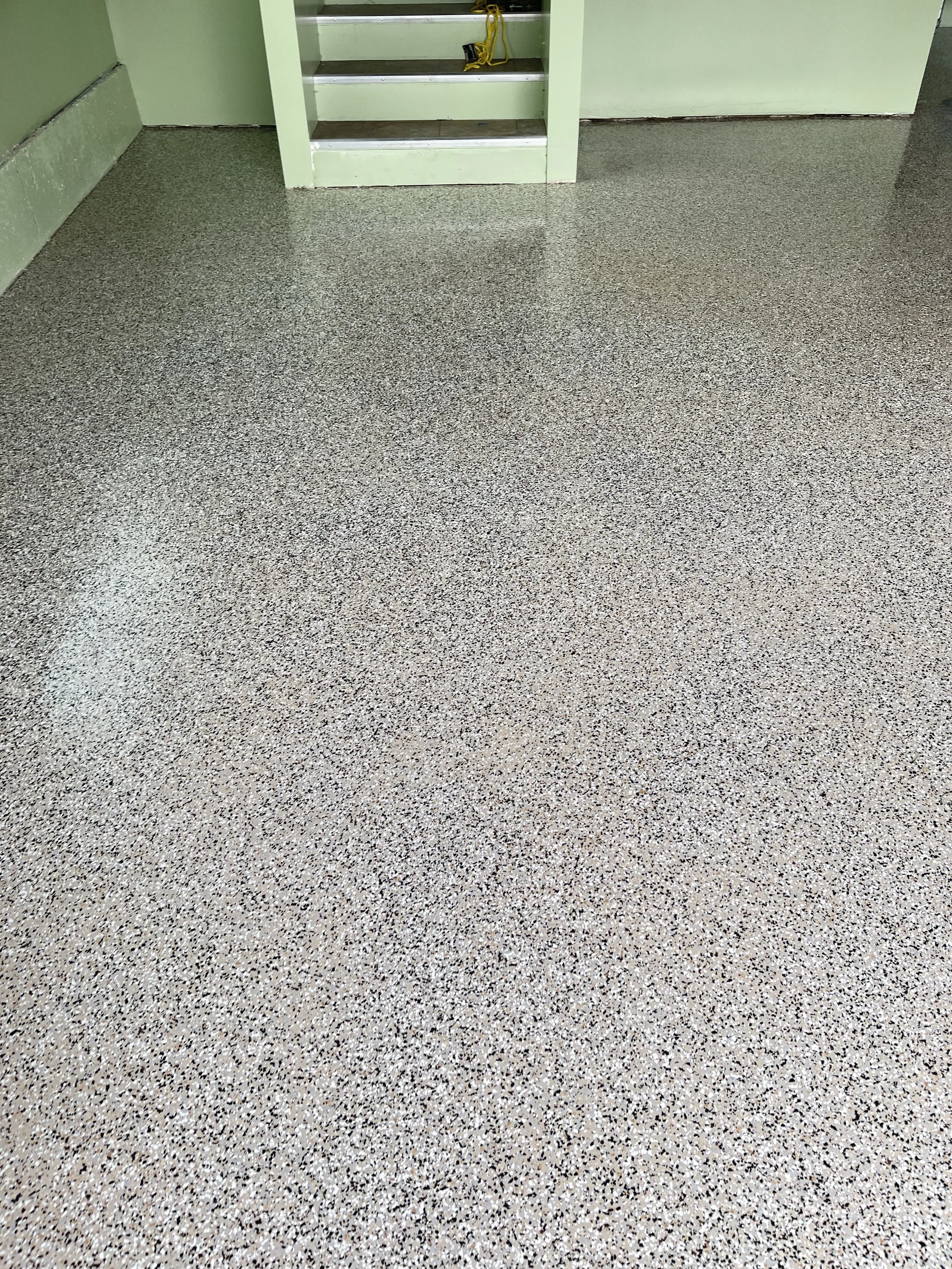 Close-up of a speckled, textured beige and black epoxy floor with a small set of light green stairs in the background.