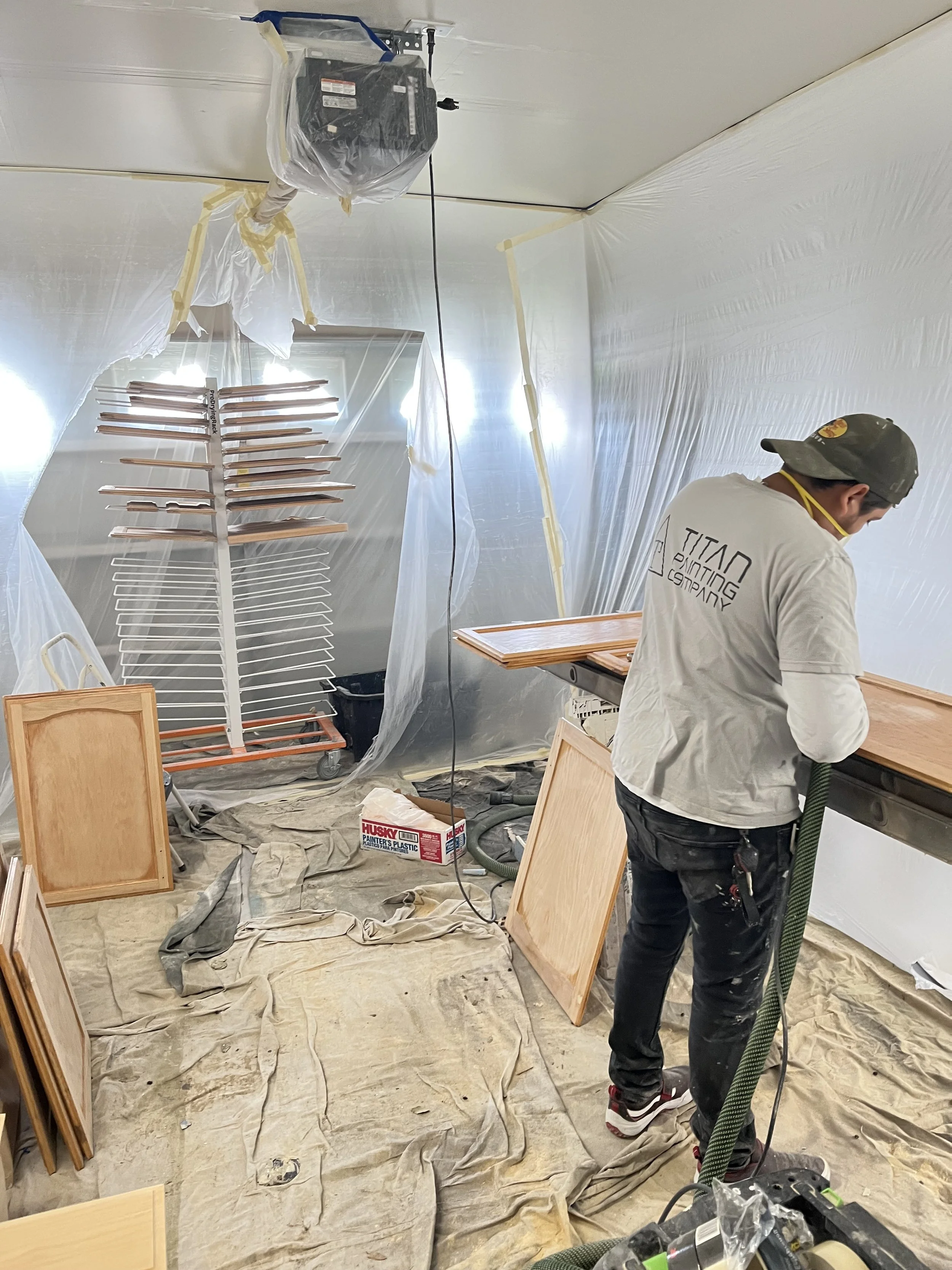 A person working on a woodworking and painting project in a room covered with plastic sheeting and drop cloths, with wooden cabinets and frames around.