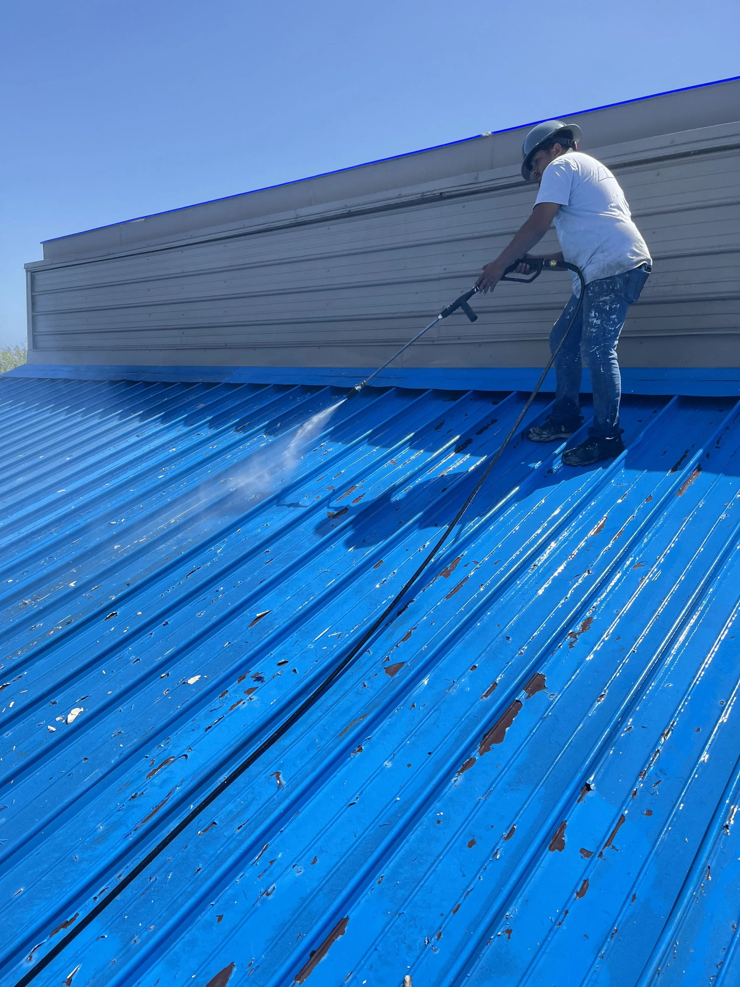Worker wearing a helmet power washing a blue metal roof.