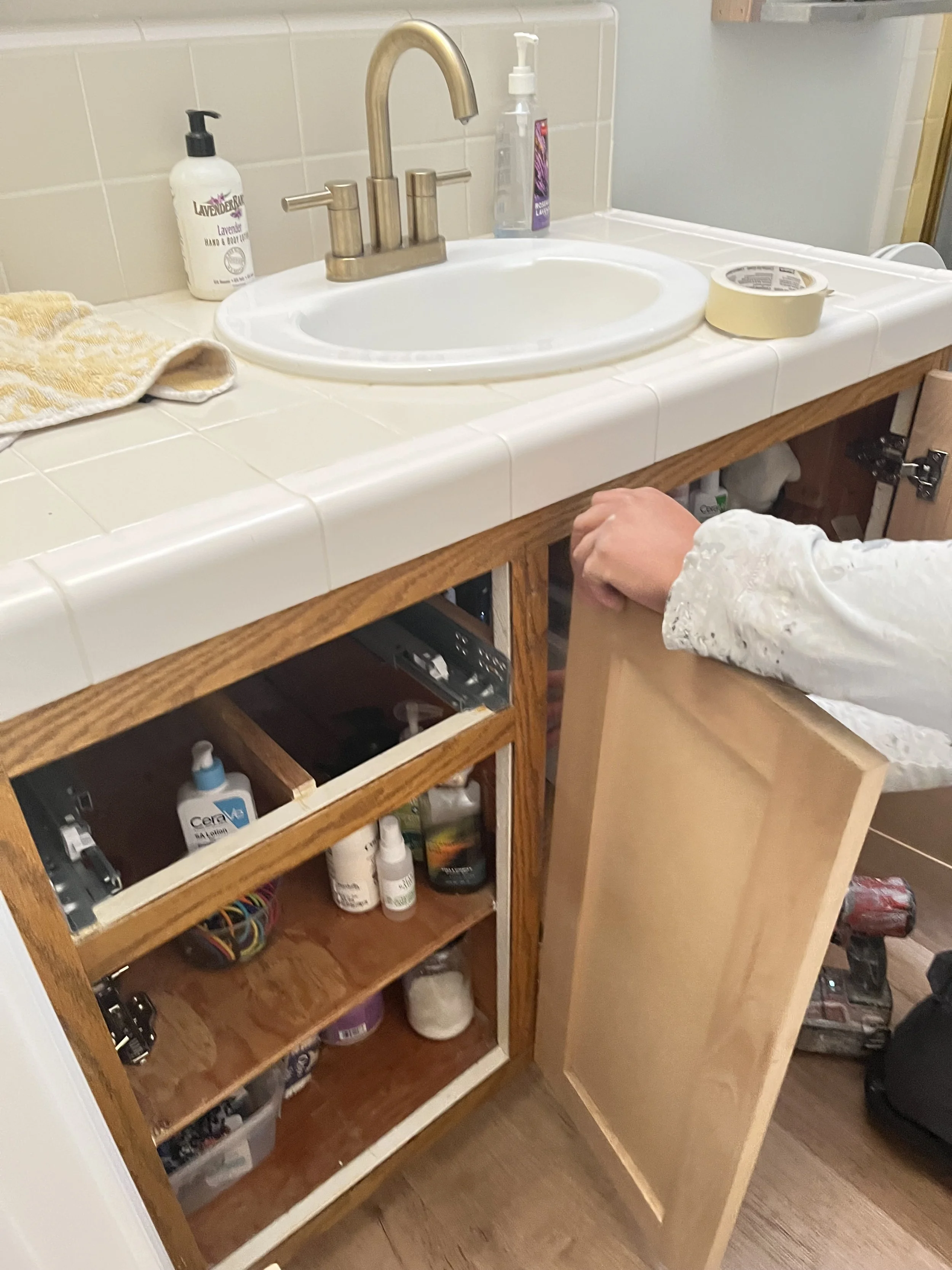 Bathroom vanity cabinet with open door, showing various toiletries, and a person holding the door open while standing in front of a white tiled countertop with a small white sink, gold faucet, hand soap, and a roll of painter's tape.