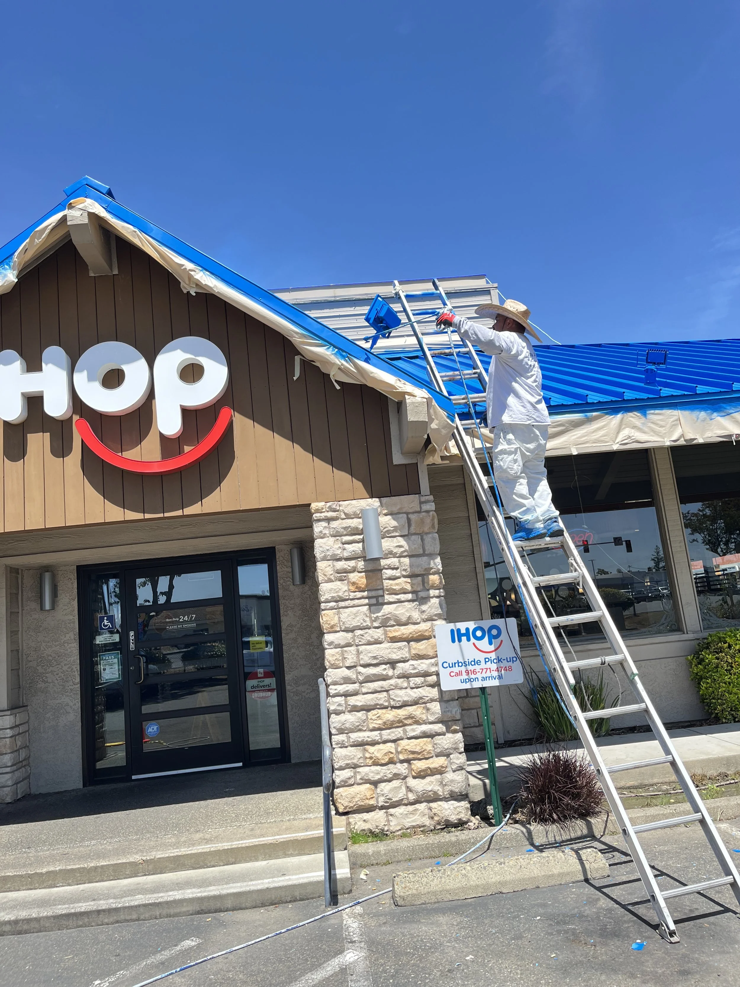 A worker painting a blue metal roof at an IHOP restaurant, standing on a ladder while wearing a white protective suit and a wide-brimmed hat, under a clear blue sky.