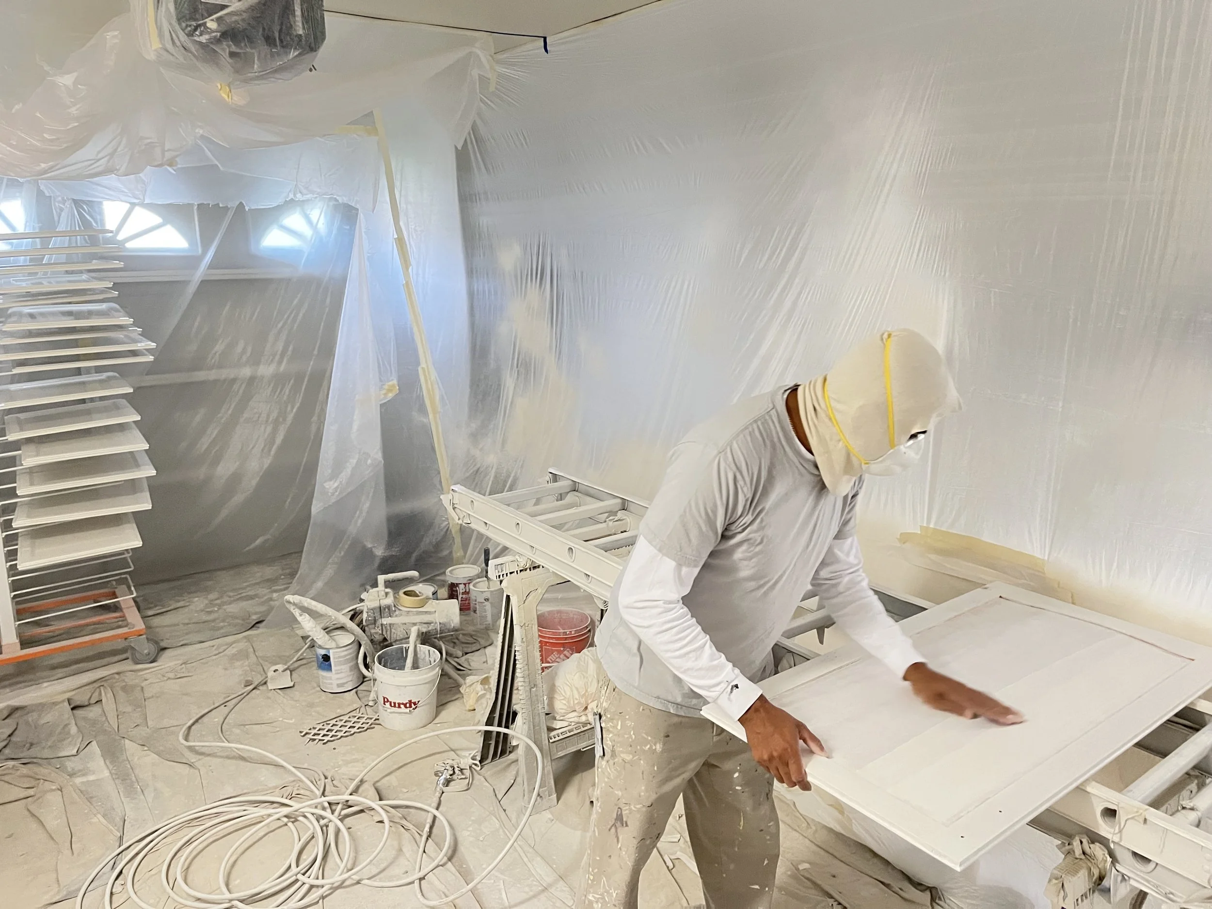 Person wearing protective clothing and face mask sanding a large white board in a room under renovation, surrounded by painting supplies and plastic-covered walls.