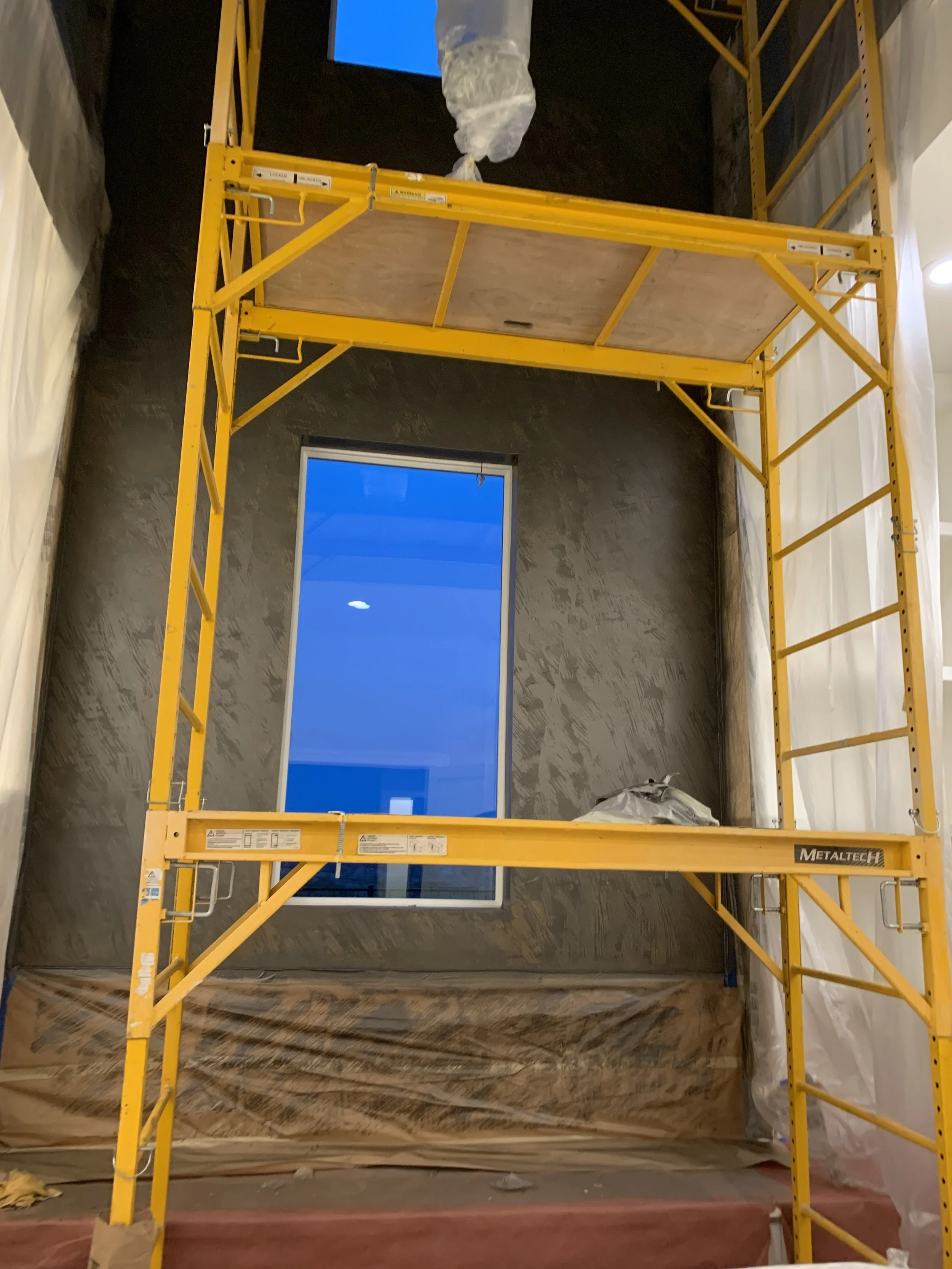 Construction scaffolding inside a room under renovation, with a window on the back wall, and plastic sheeting covering parts of the room.