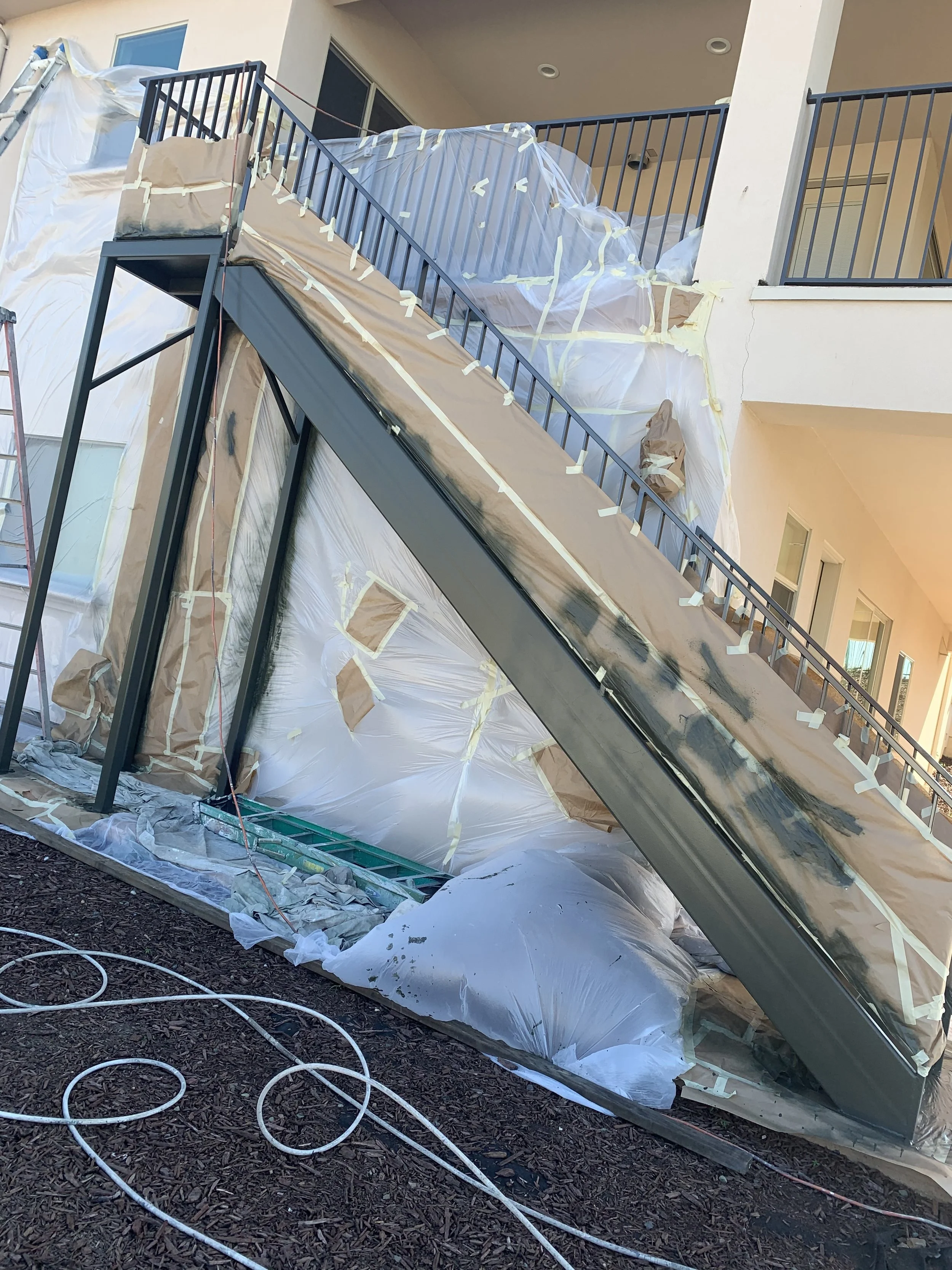 Construction site featuring a staircase and balcony, covered with plastic and paper for painting or renovation, with tools and cords on the ground.