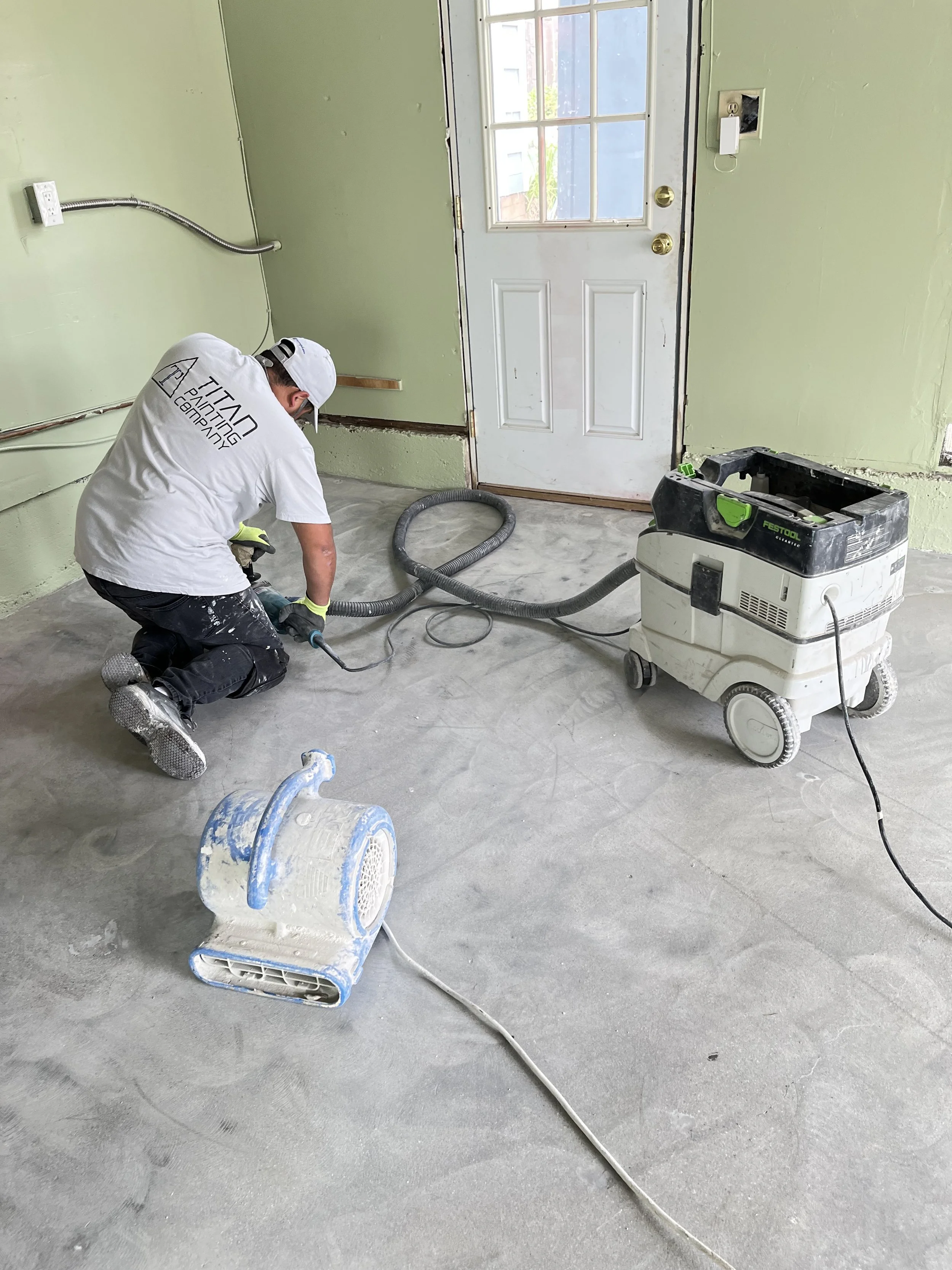 A worker kneeling on the floor using a concrete grinder machine in a room under renovation, with another smaller blue grinder nearby and a hose connected to a vacuum system.