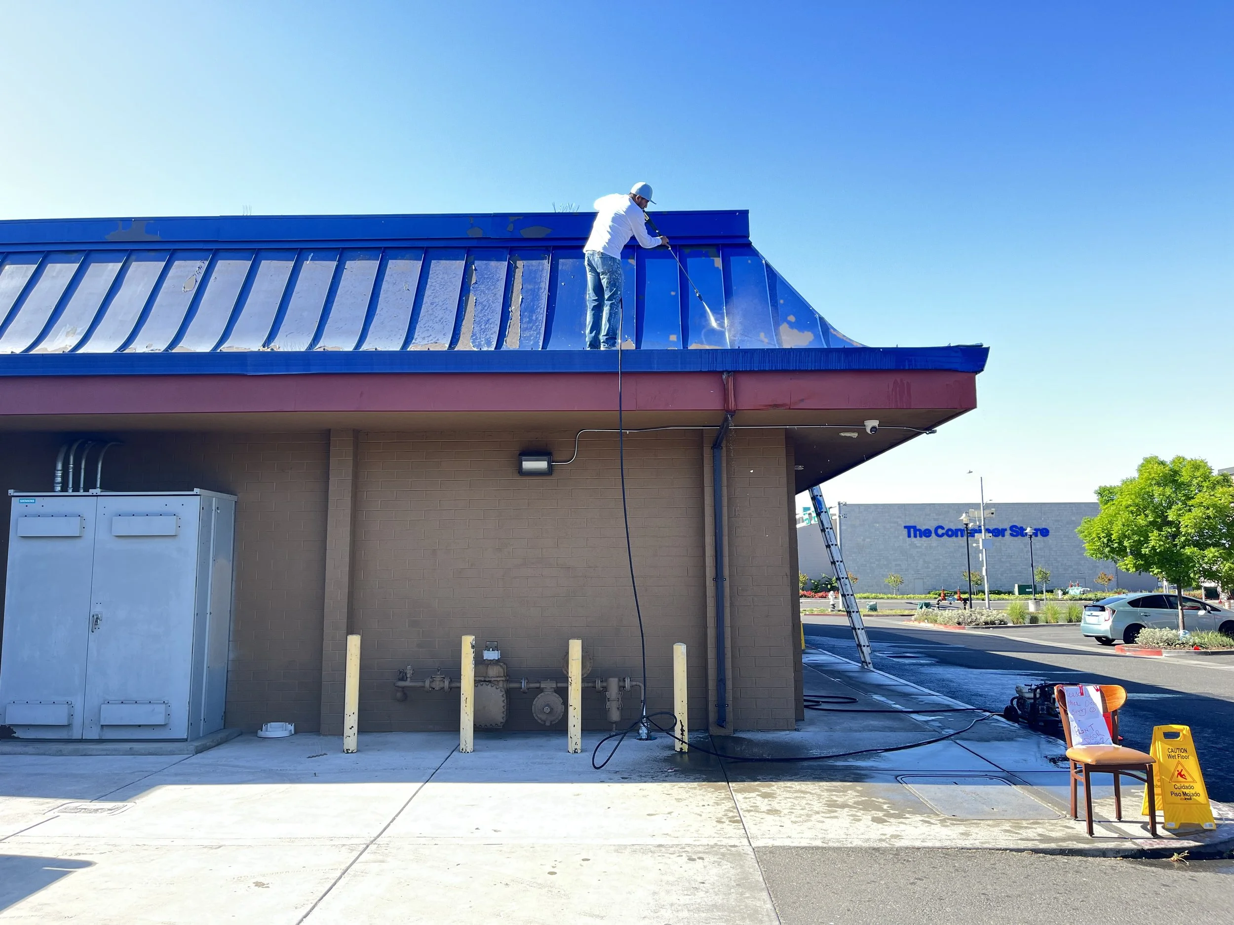 A worker power washing the roof of a commercial building in a shopping center parking lot, with a ladder leaning against the building and a chair with a caution wet floor sign nearby.