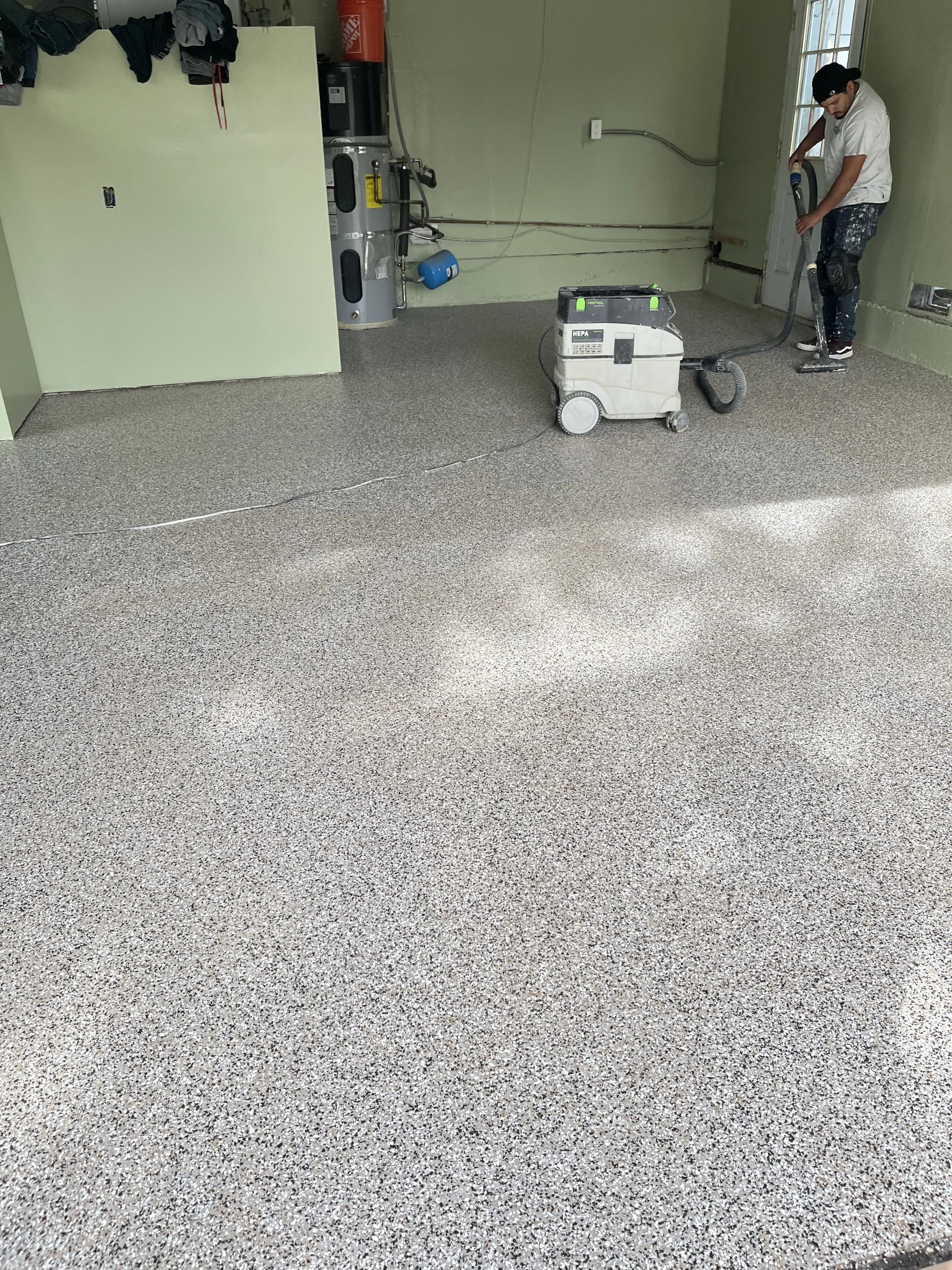 A man is vacuuming a speckled beige and gray epoxy floor in a room with green walls and a window. There is a water heater, water dispenser, and some laundry hanging on the wall.