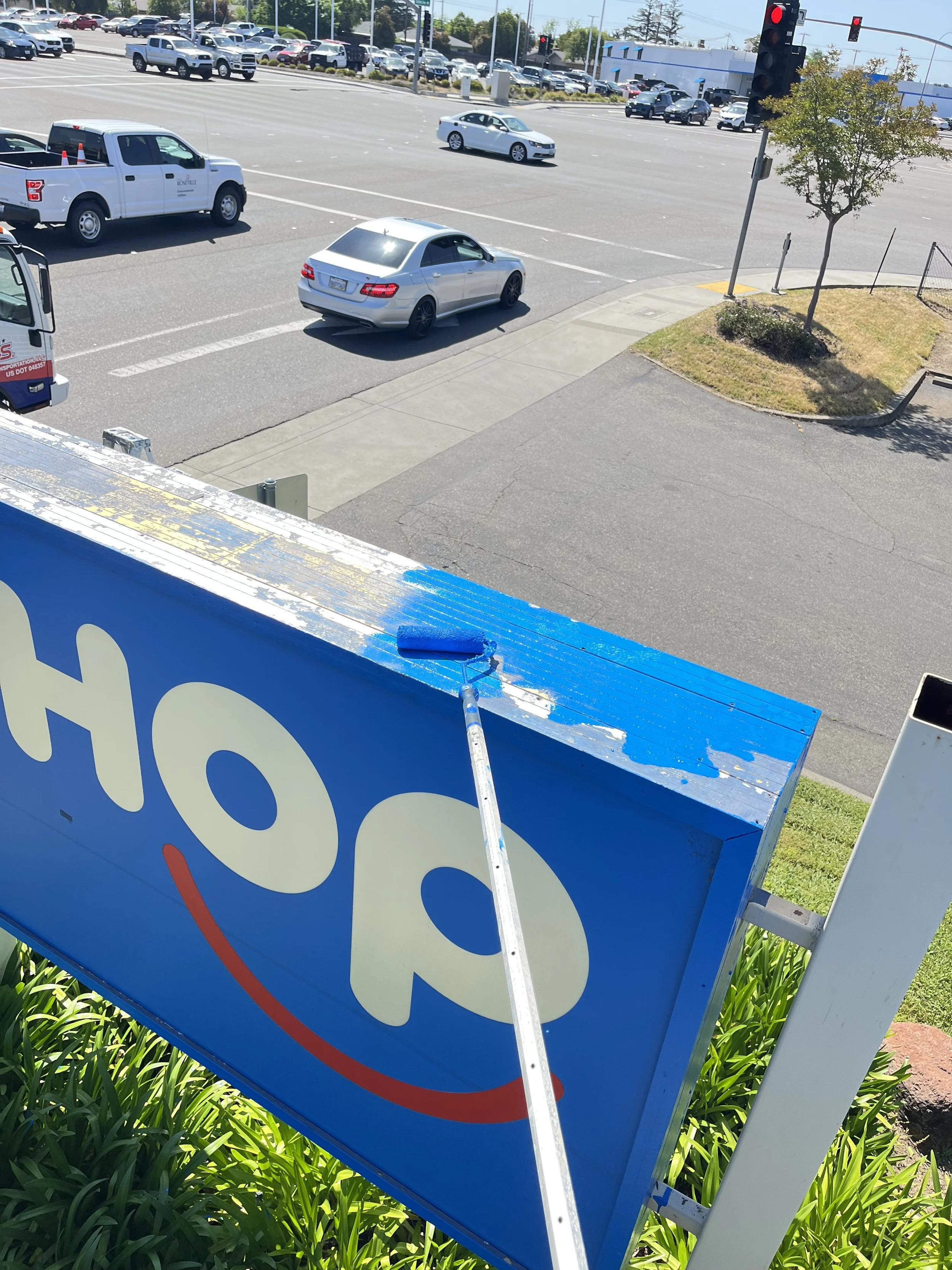 Photo taken from an elevated perspective showing a blue and white sign being painted with blue paint, a sidewalk, a small landscaped island with a tree, and a busy street intersection with several cars and trucks waiting at traffic lights.
