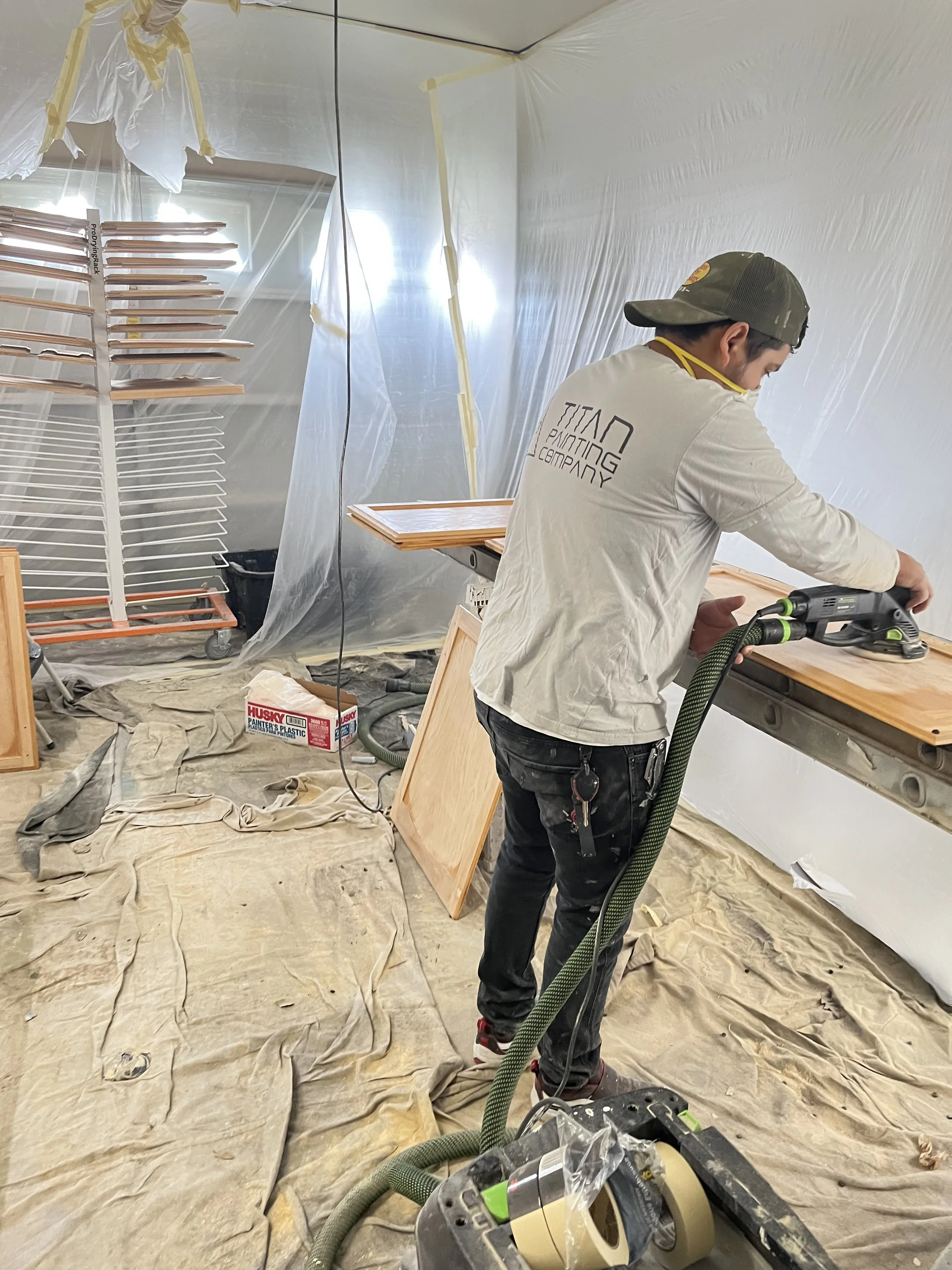 A person sanding a wooden surface in a makeshift painting booth with plastic sheeting walls, construction supplies, and towels on the floor.