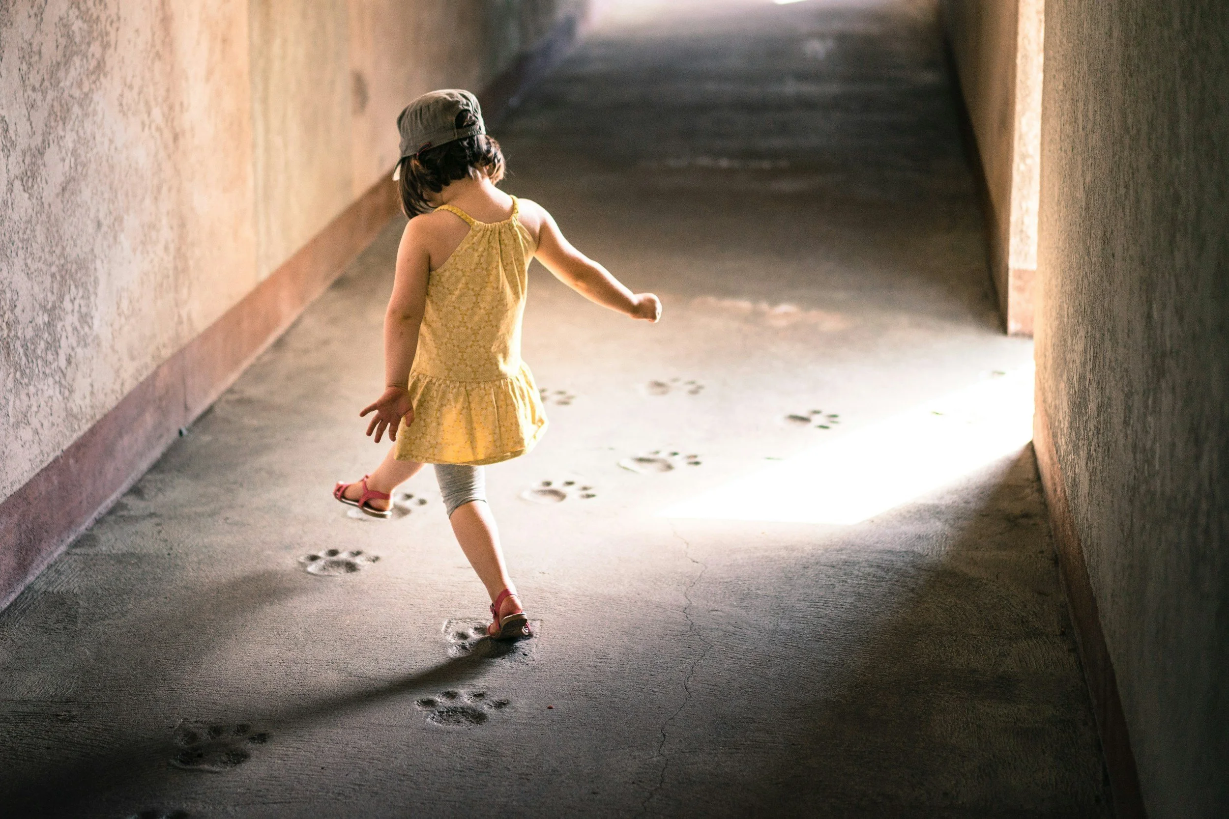 A young girl wearing a yellow dress, gray leggings, and red sandals is walking through a dimly lit corridor, leaving paw prints on the ground.