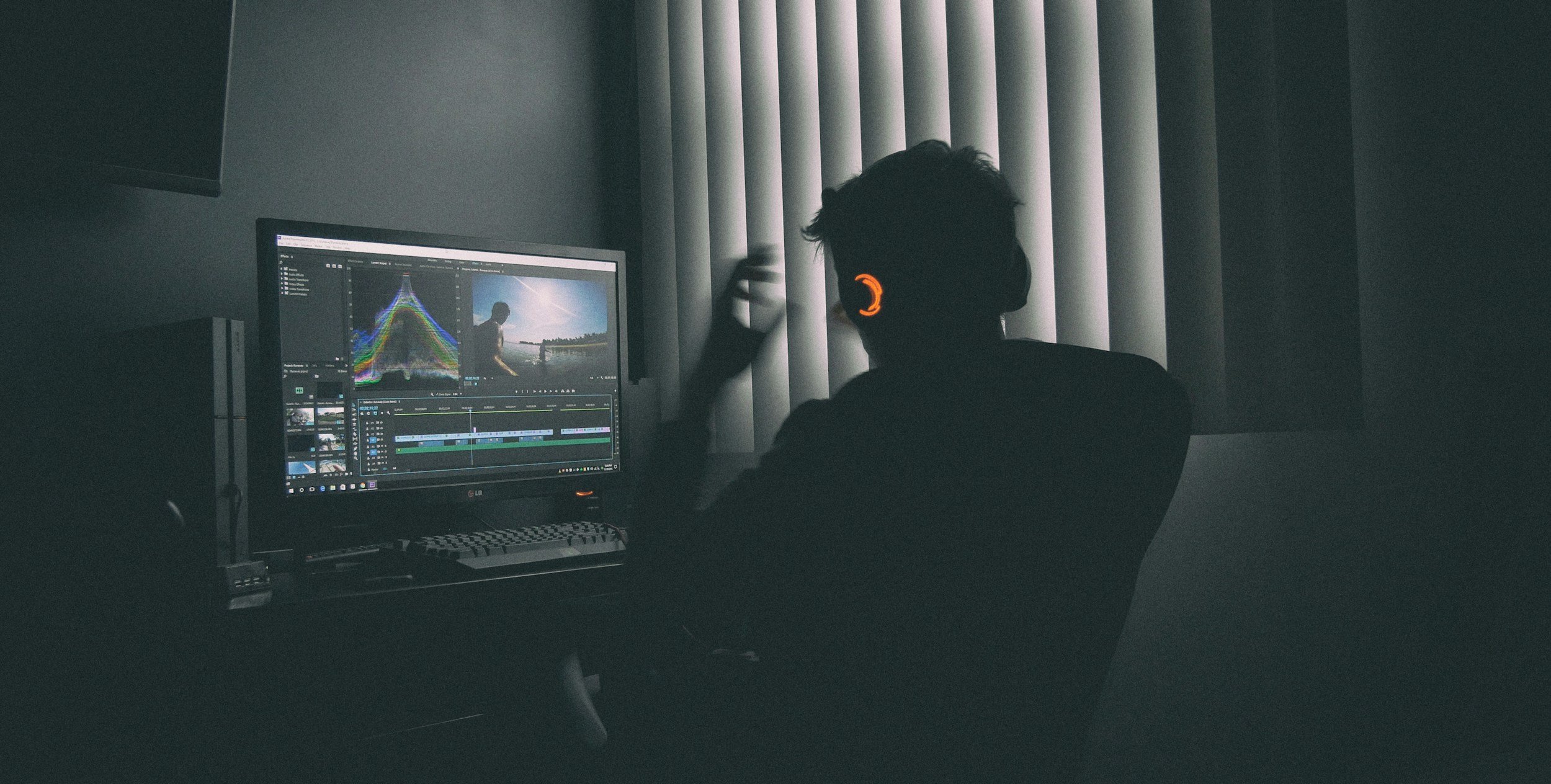 Person working on video editing at a computer in a dark room with blinds.