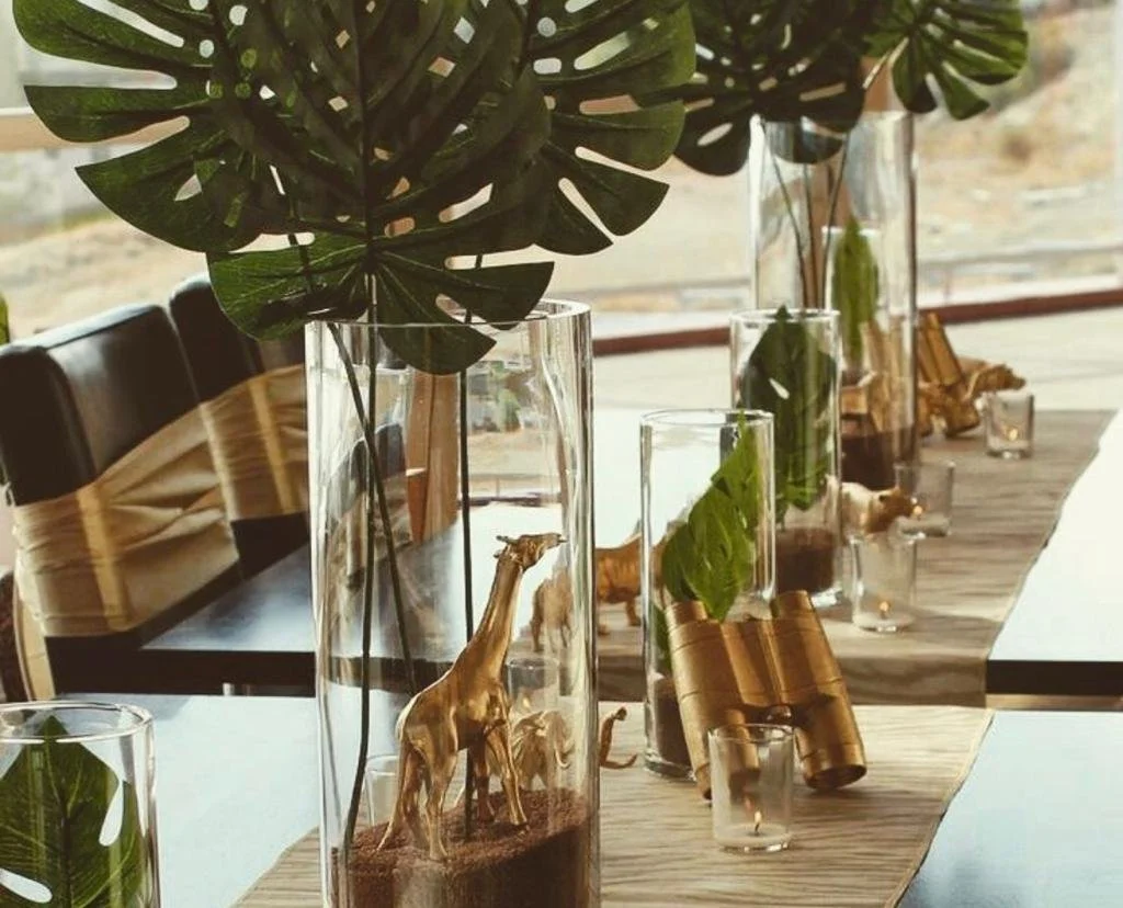 Table centerpiece featuring large green leaves in tall glass vases with gold animal figurines and small candles.