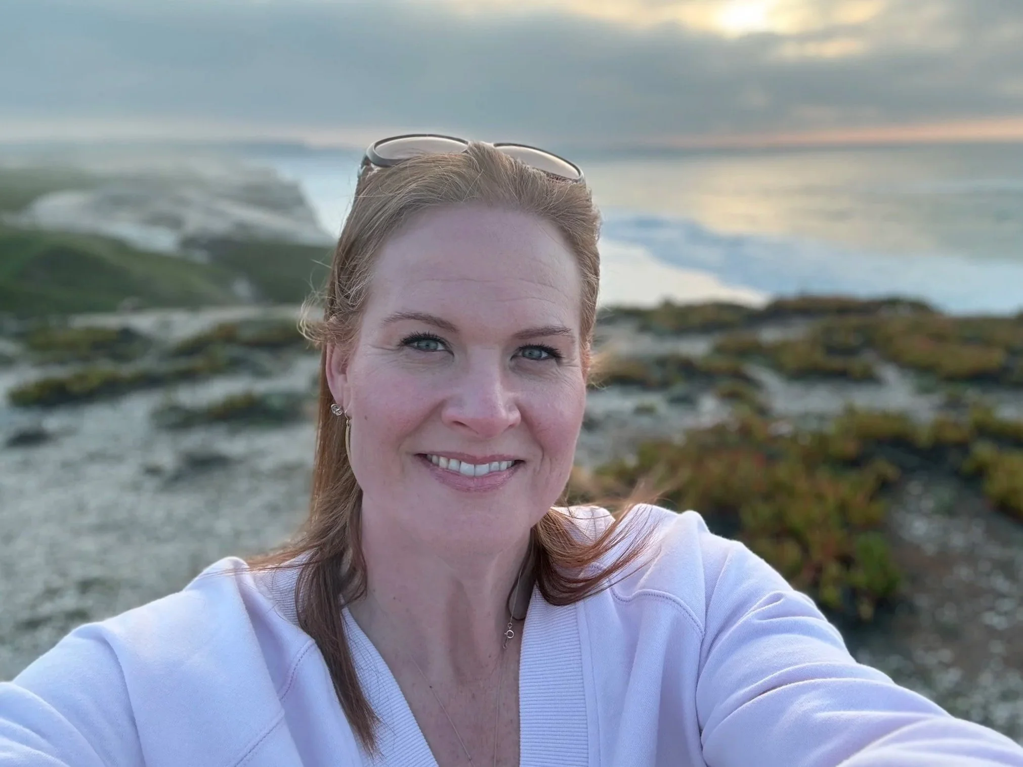 A woman with light skin and auburn hair wearing a white top, sunglasses on her head, and earrings, smiling at the camera during sunset or sunrise on a beach with rocks and seaweed in the background.