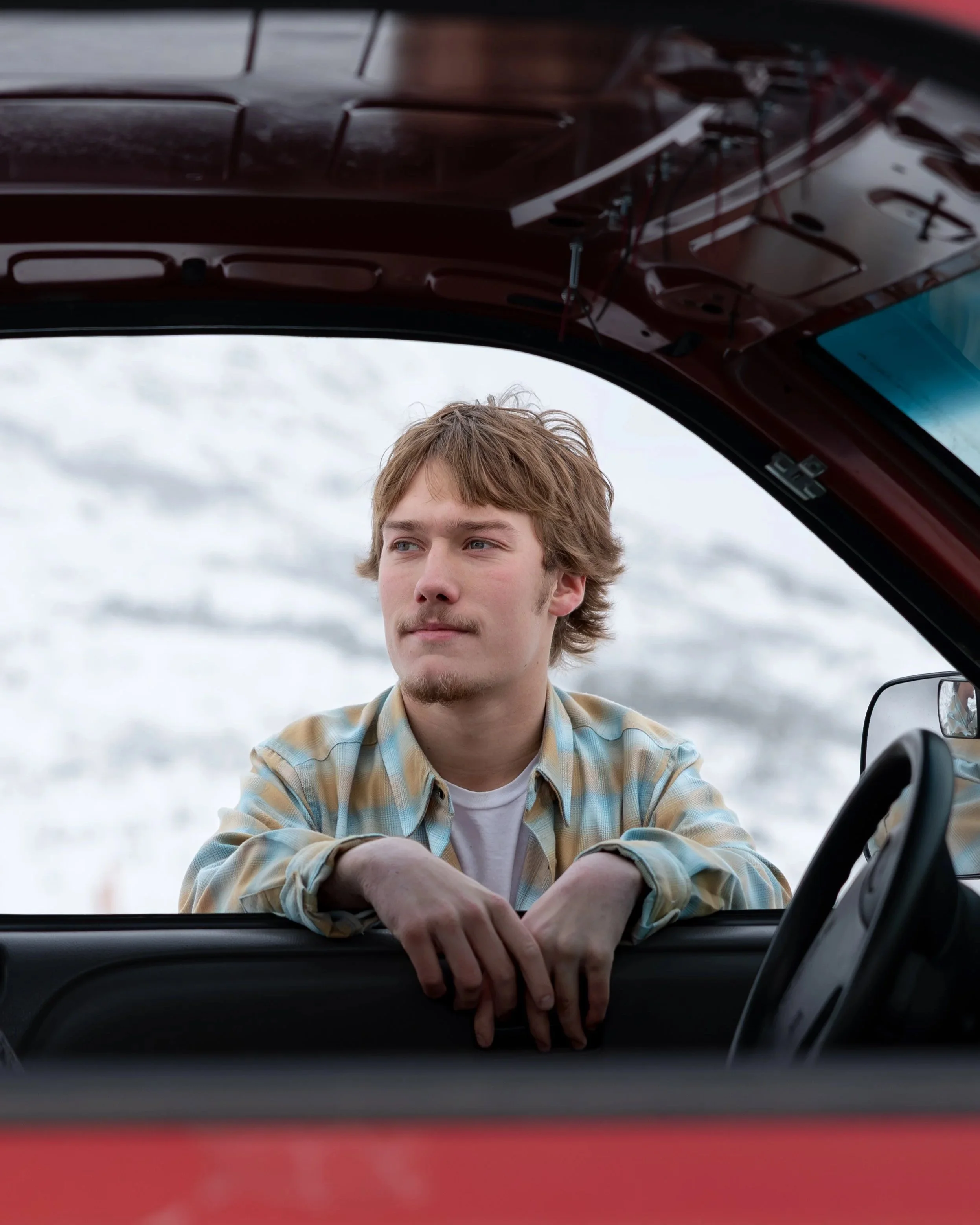 A young man with light brown hair and a beard, wearing a plaid shirt, leaning on the open window of a red vehicle, looking thoughtfully into the distance, with an overcast sky in the background.