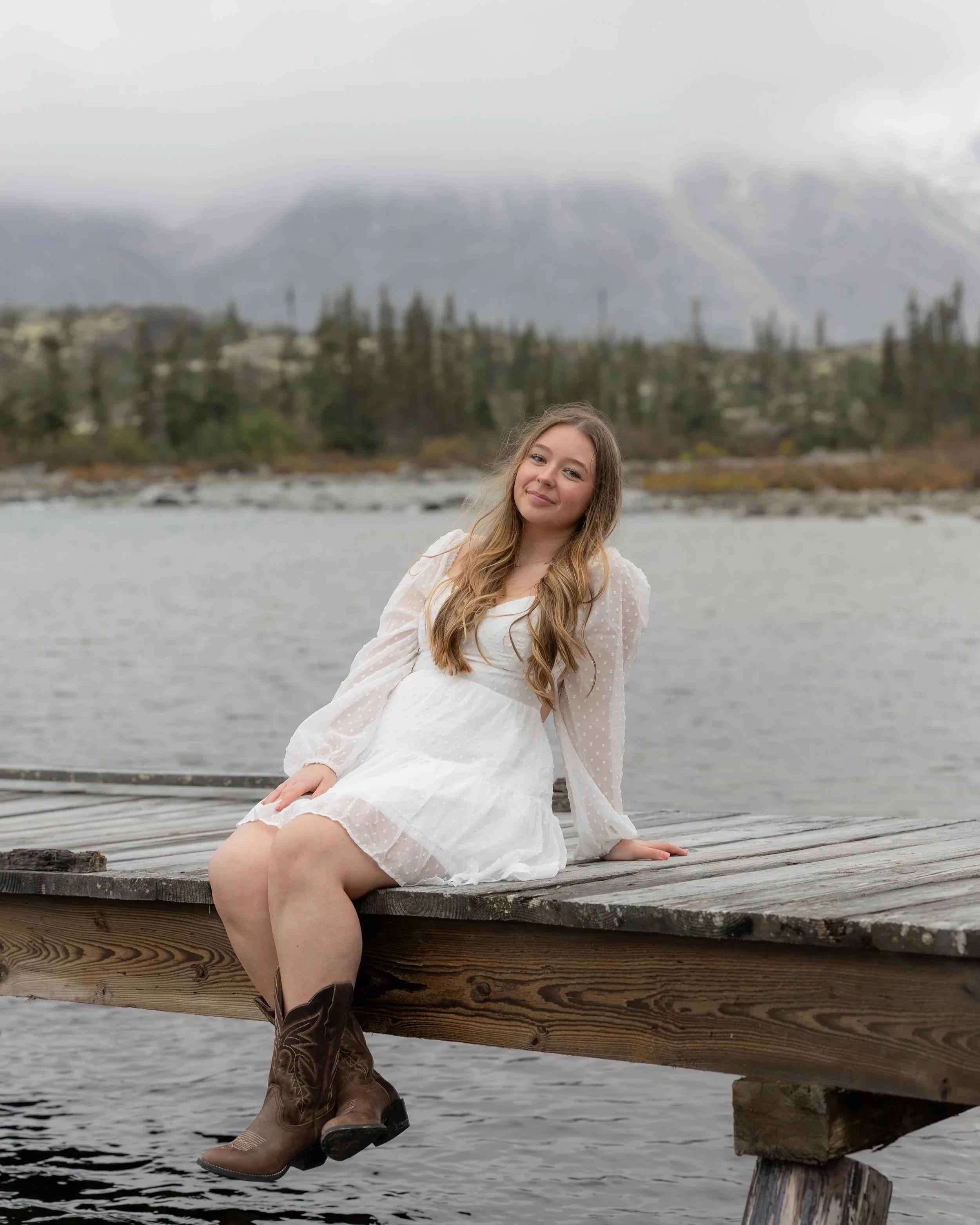 A young woman in a white dress and cowboy boots sitting on a wooden dock by a lake, with mountains and trees in the background.