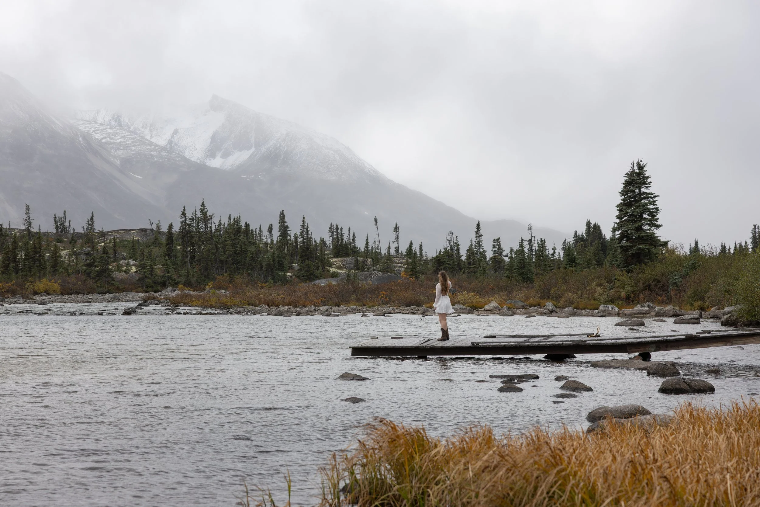 A woman in a white dress and boots standing on a wooden dock over a lake, with trees, rocky shoreline, and snow-capped mountains in the background under cloudy skies.