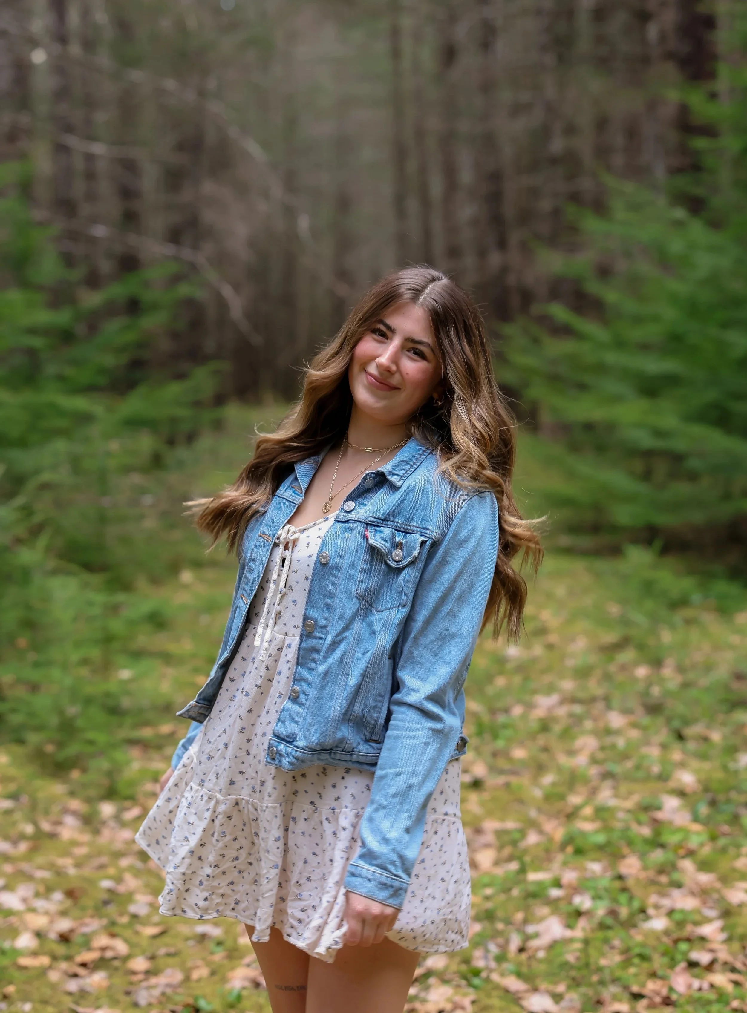 Eliza Myers, a young woman with long wavy hair, smiles and stands in a lush green forest, wearing a white dress with small flowers and a light blue denim jacket.