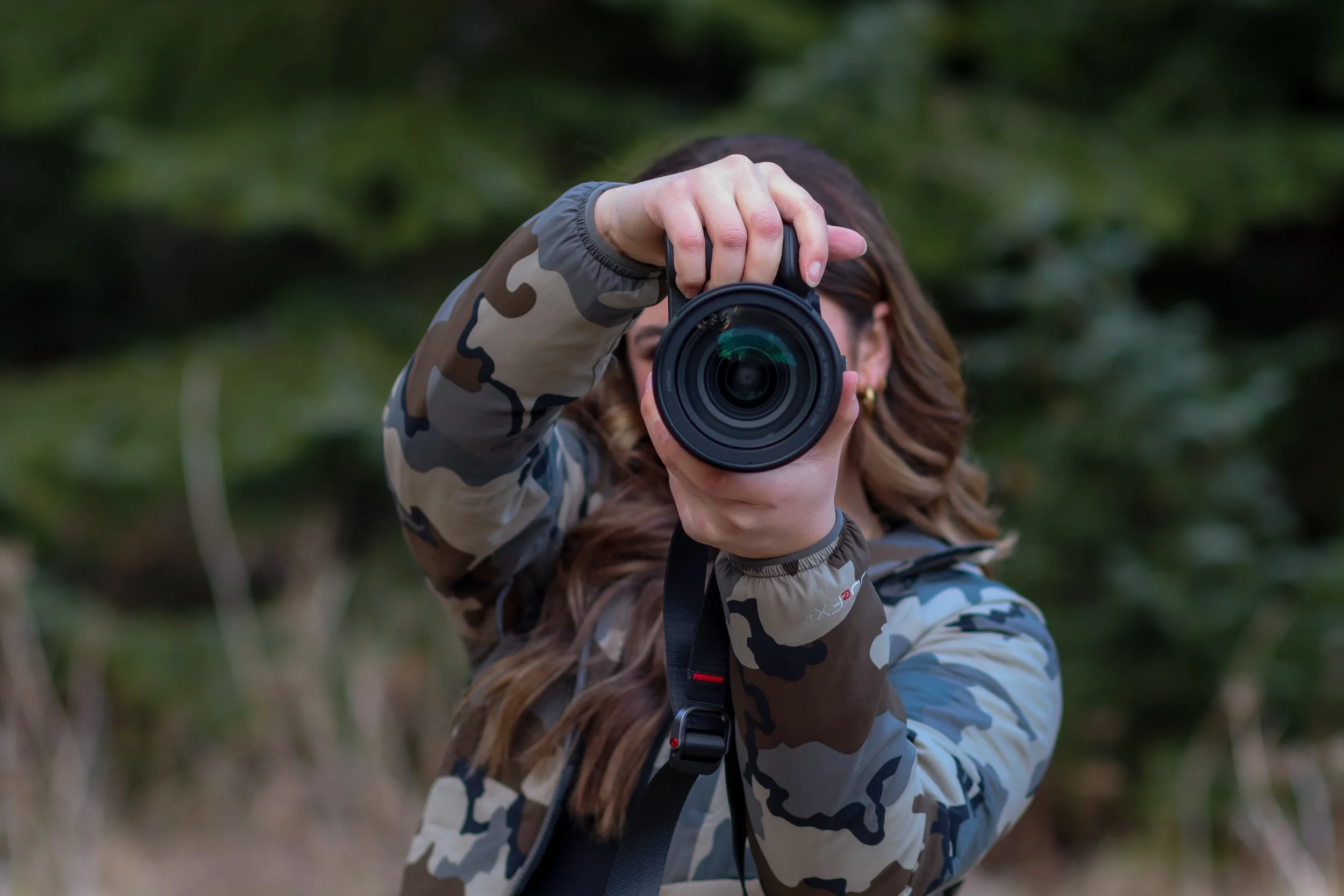 Person holding a camera, taking a photo outdoors with a blurred green foliage background.