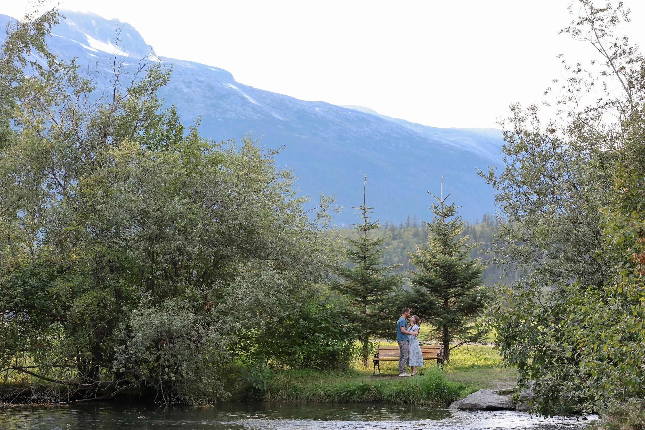 A couple standing and embracing near a river with trees and mountains in the background.