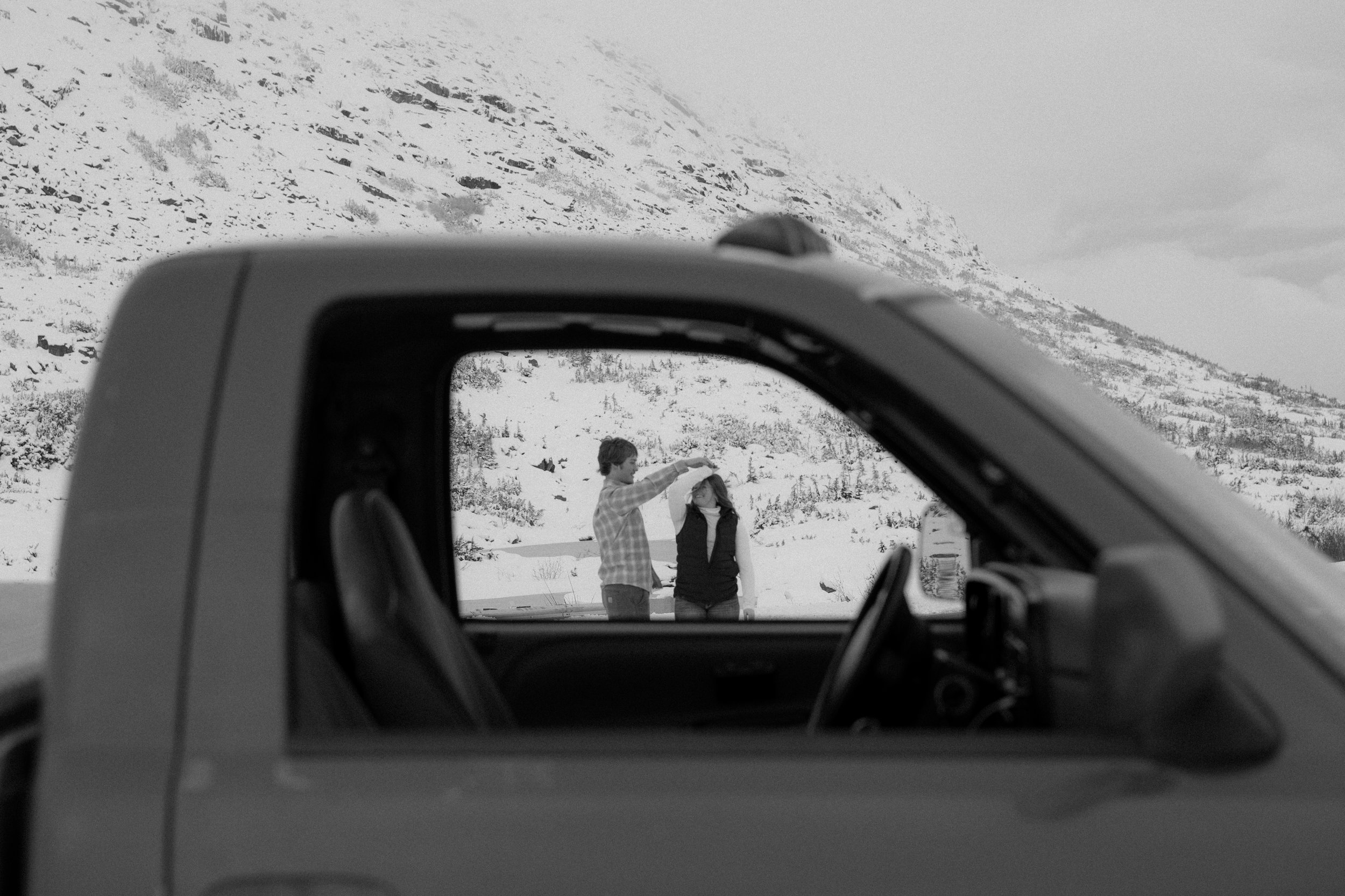 A person in a checkered shirt is using their hand to place a necklace on a woman standing outside in a snowy landscape, with mountains in the background, viewed through the window of a vehicle.