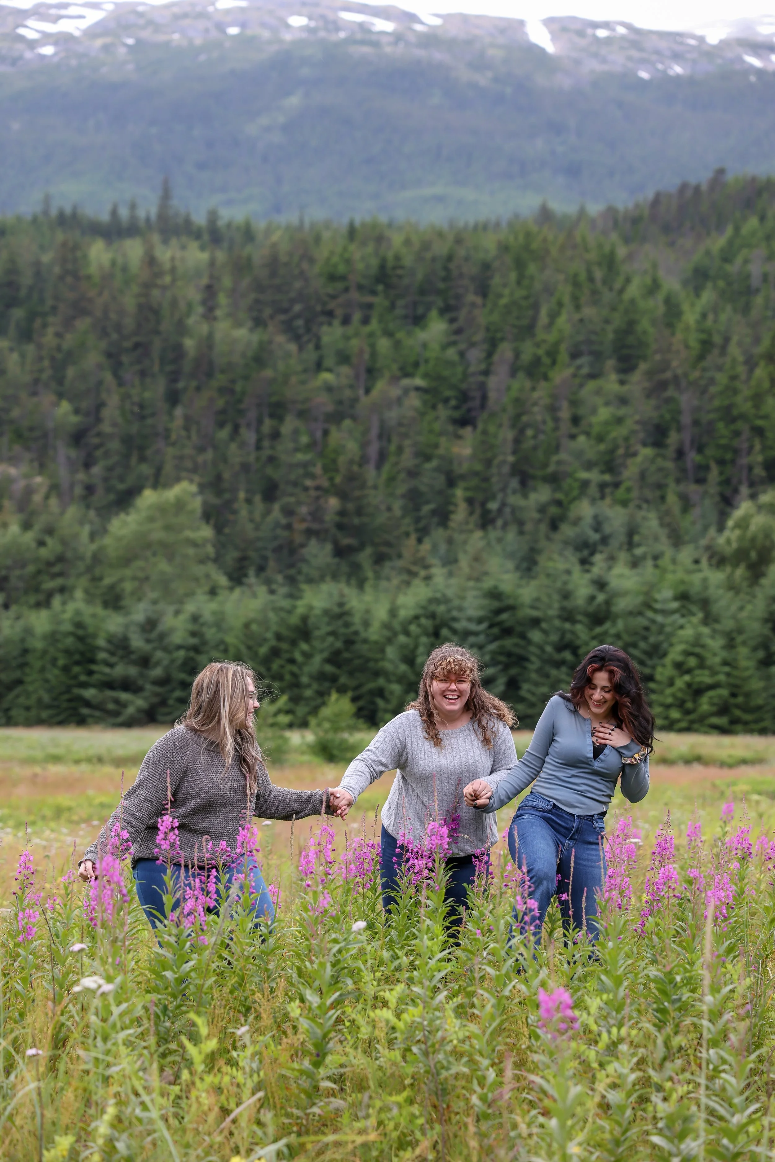 Three women holding hands and running through a field of pink flowers with a forest and mountain in the background.