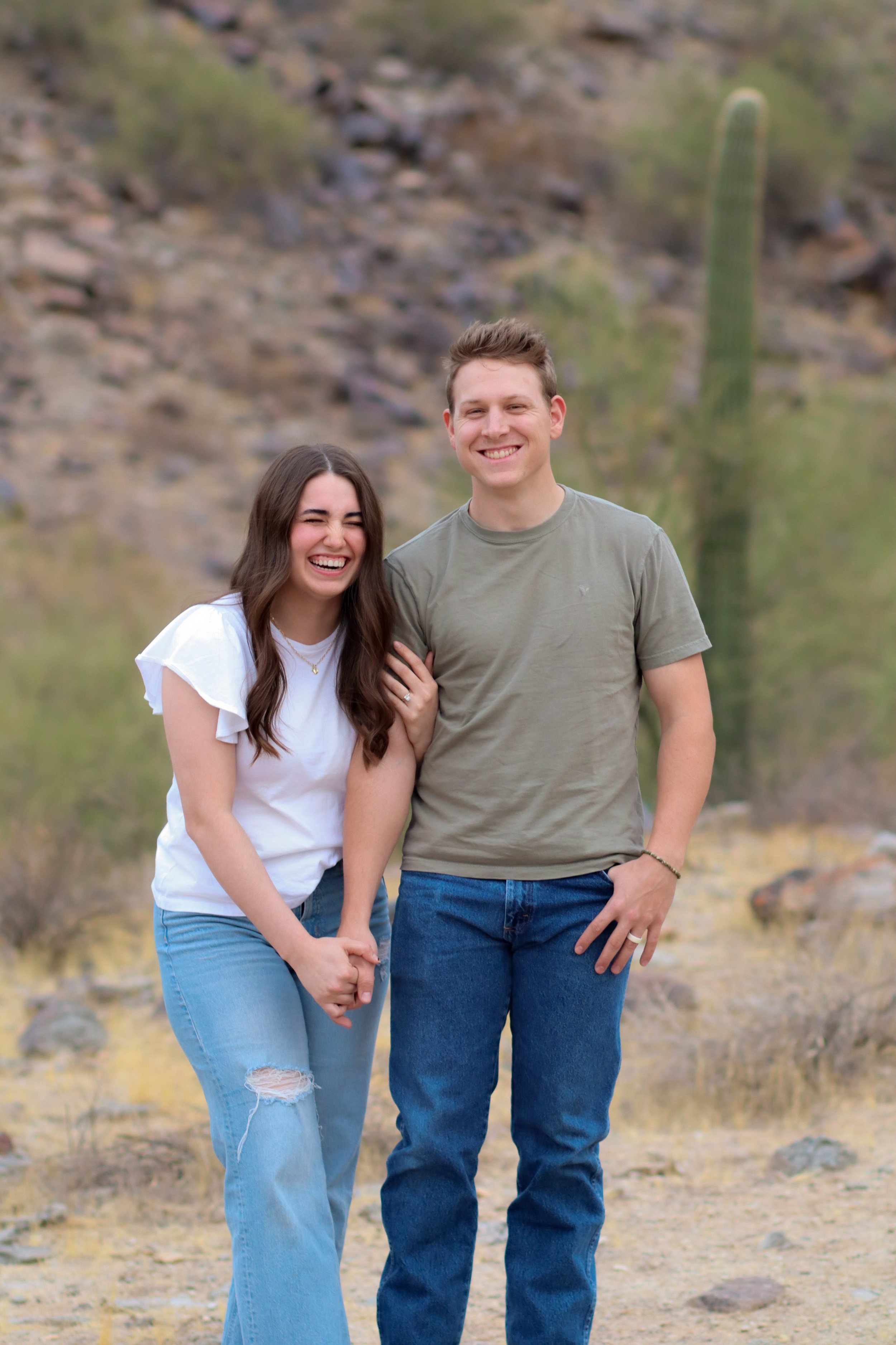 A young couple is outdoors in a desert landscape, smiling and laughing together. The woman has long dark hair, is wearing a white t-shirt and ripped jeans, and is holding the man's hand. The man has short light brown hair, is wearing a gray t-shirt and blue jeans. There is a large cactus and rocky hills in the background.