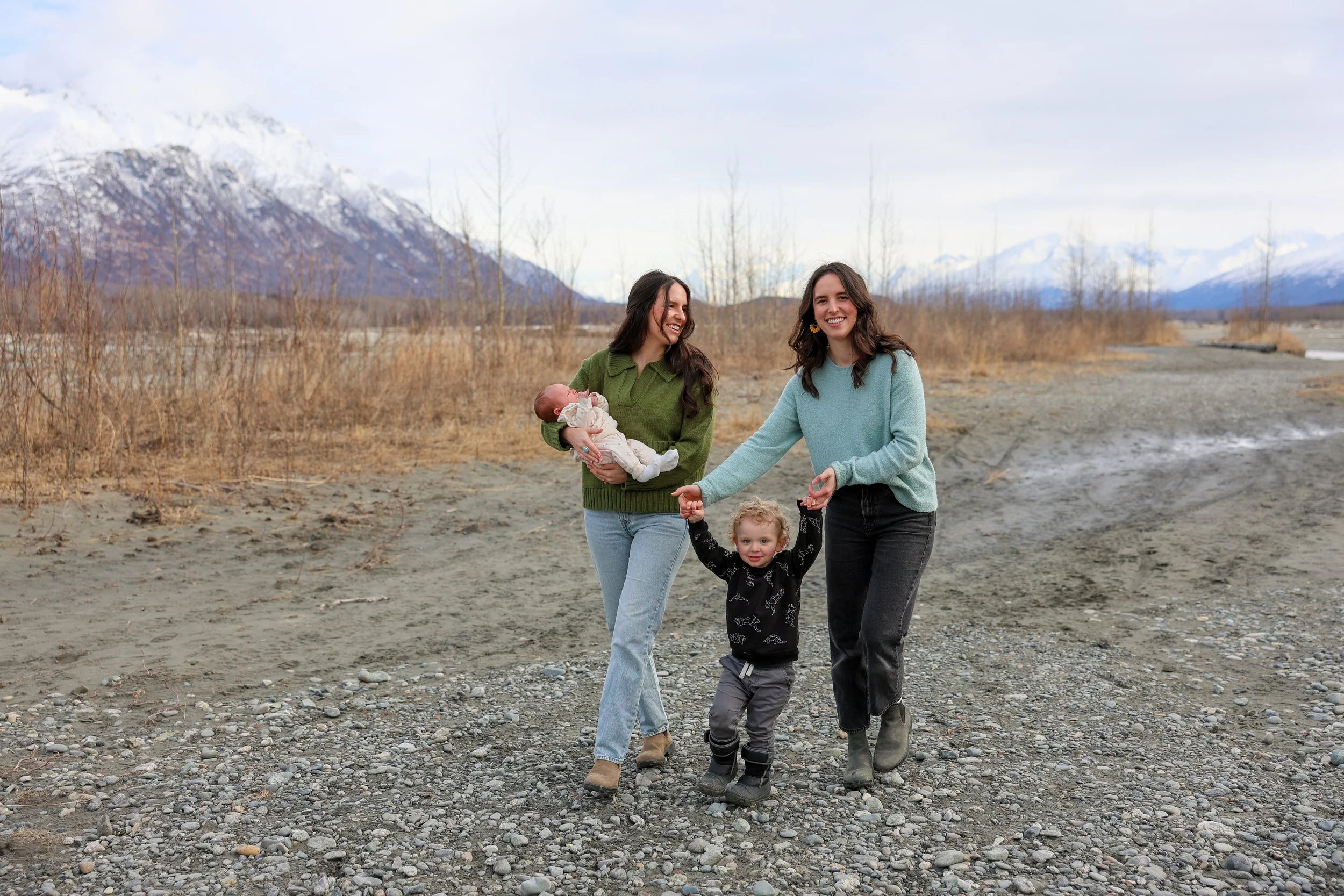 Two women and a boy walk along a rocky riverbank, with mountains in the background. One woman holds a baby, and the boy is holding both women's hands.