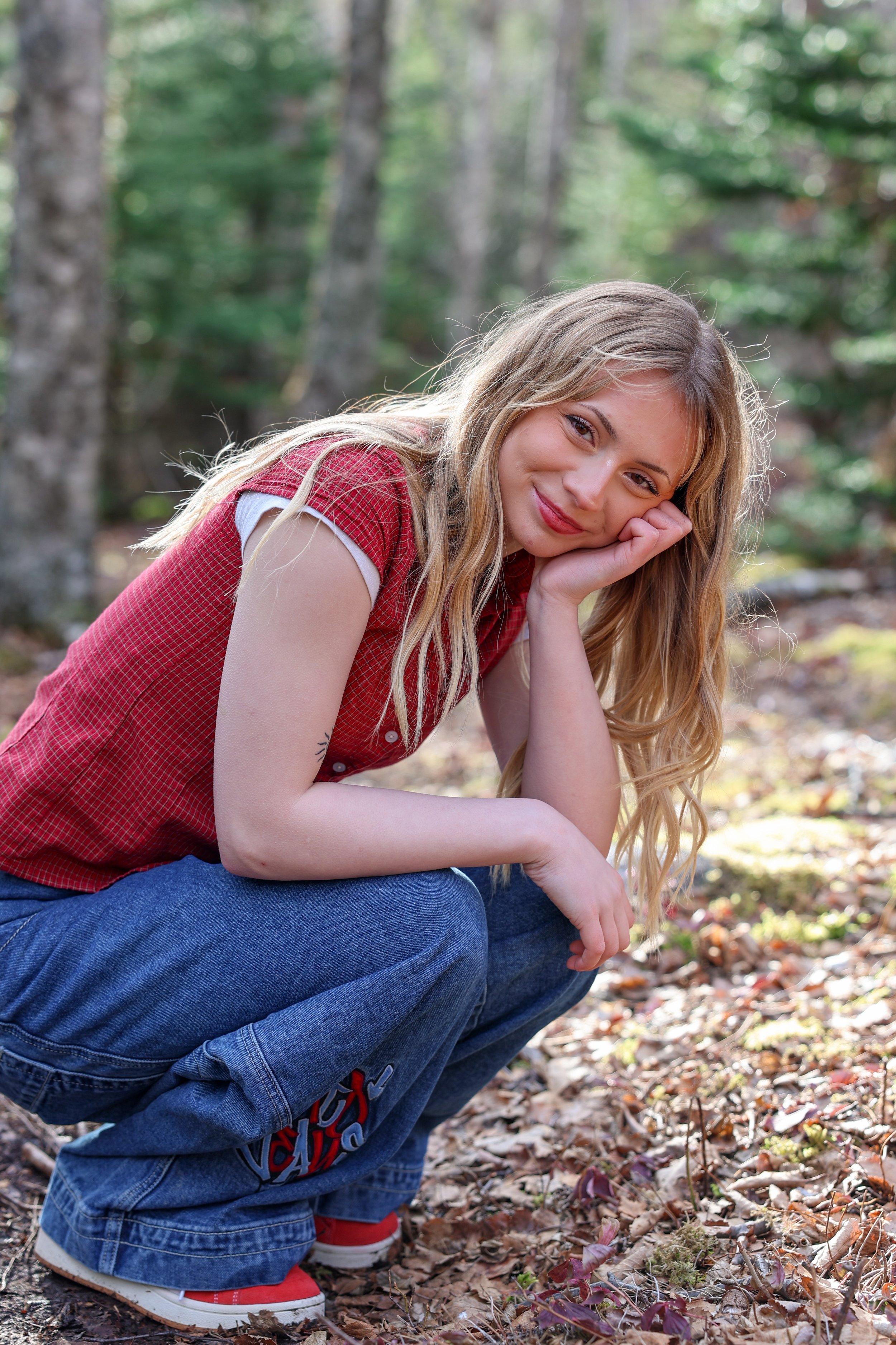 Young woman crouching outdoors in a forest, resting her chin on her hand and smiling.