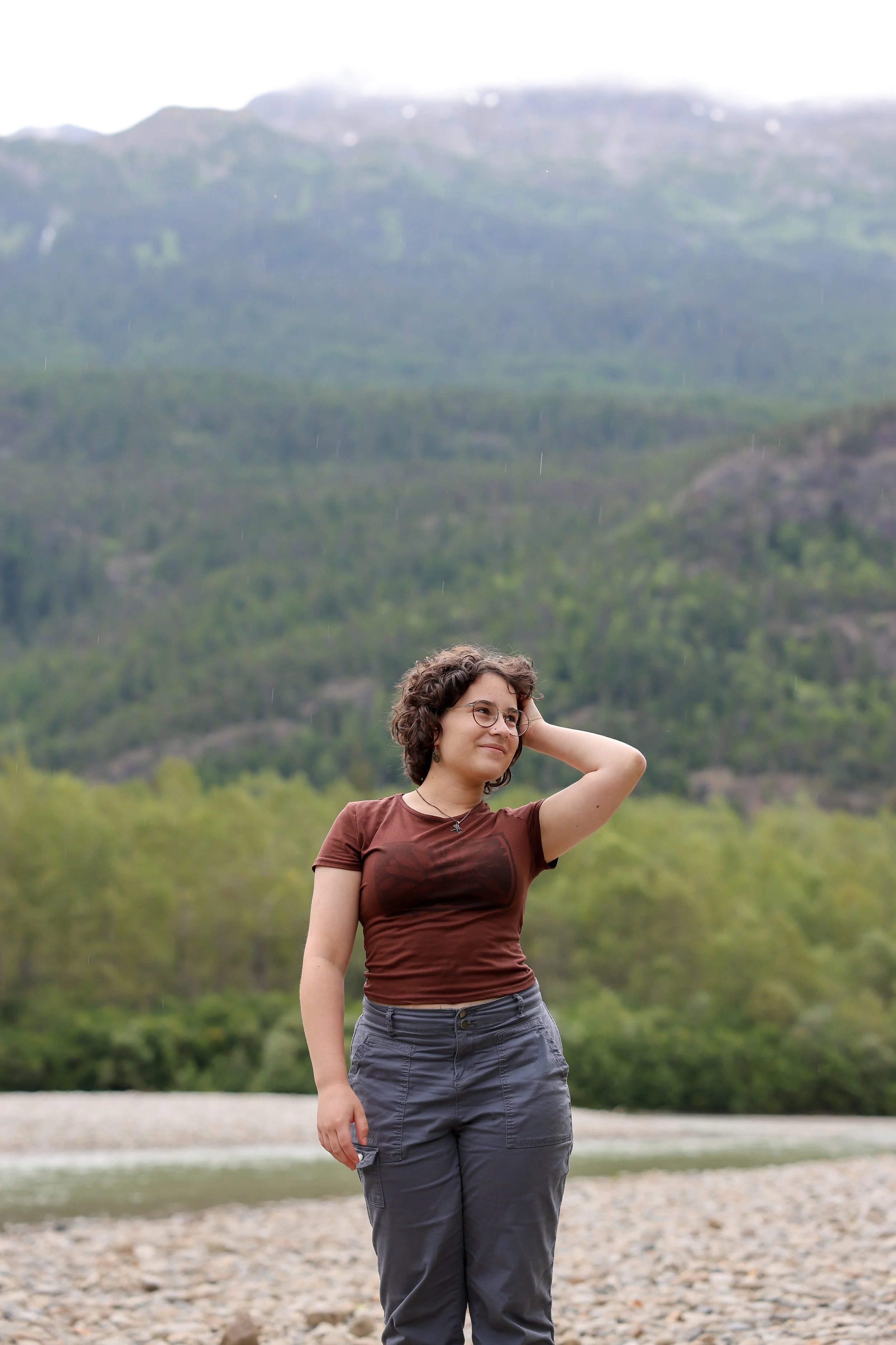 A young woman with curly hair, glasses, and a brown t-shirt standing outdoors on a rocky riverbank, with lush green trees and mountainous terrain in the background on a rainy day.