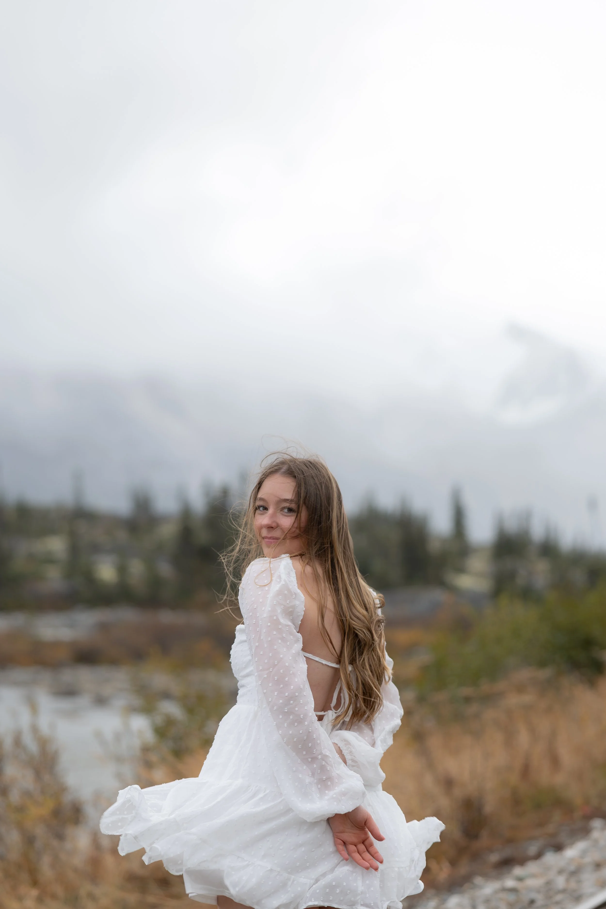 A young girl wearing a white dress standing outdoors with a cloudy sky and trees in the background.