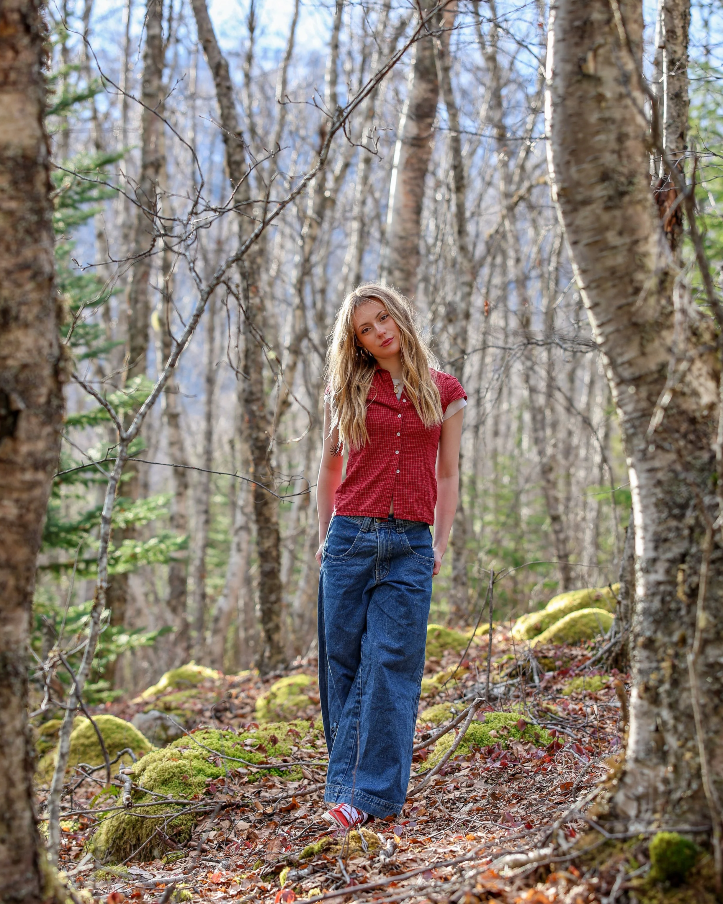A young woman with long blonde hair standing alone in a sparse forest with leafless trees, wearing a red short-sleeved shirt, baggy blue jeans, and red shoes, with a relaxed expression.