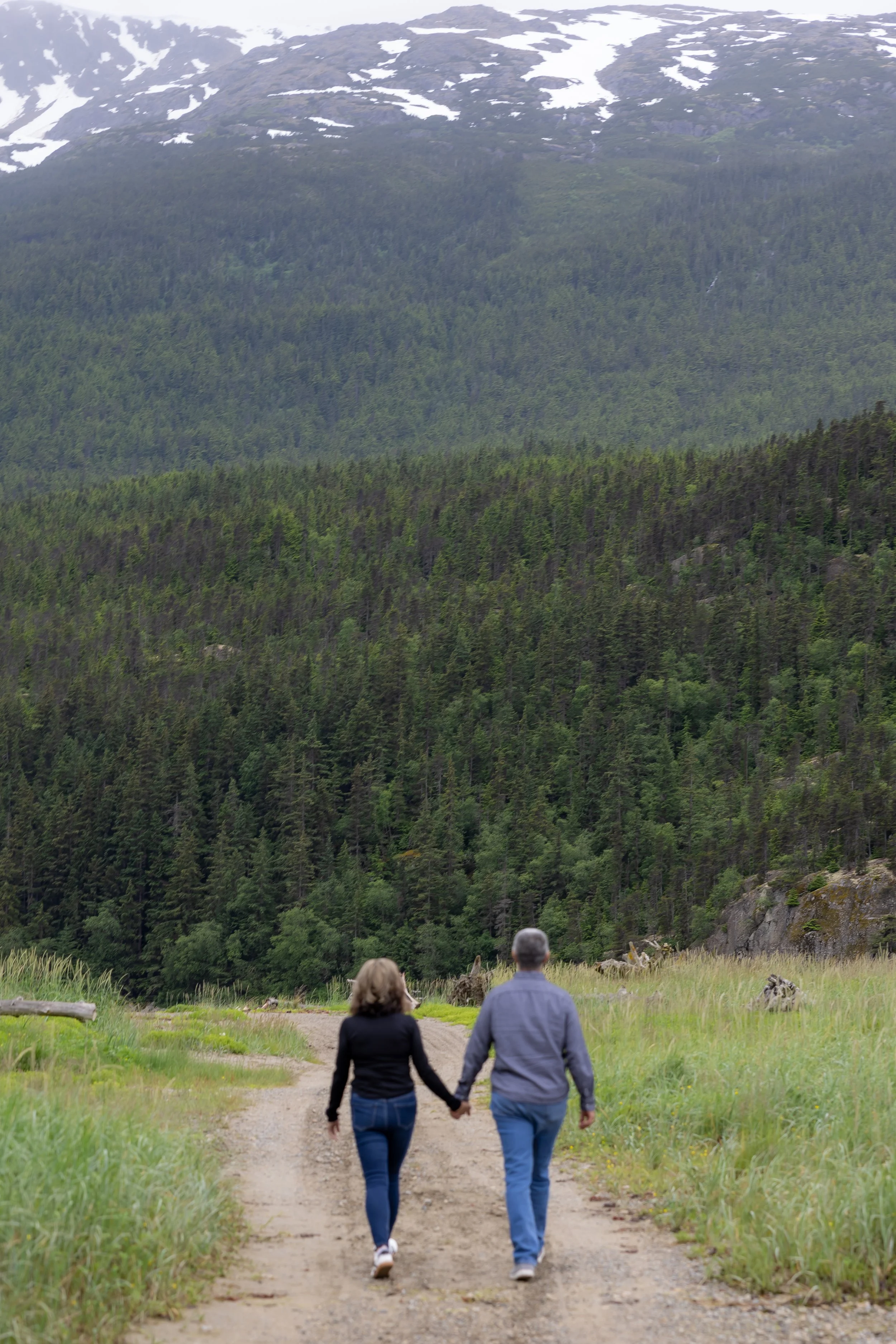 A couple walking hand in hand on a dirt trail through a green field with a forested mountain in the background.