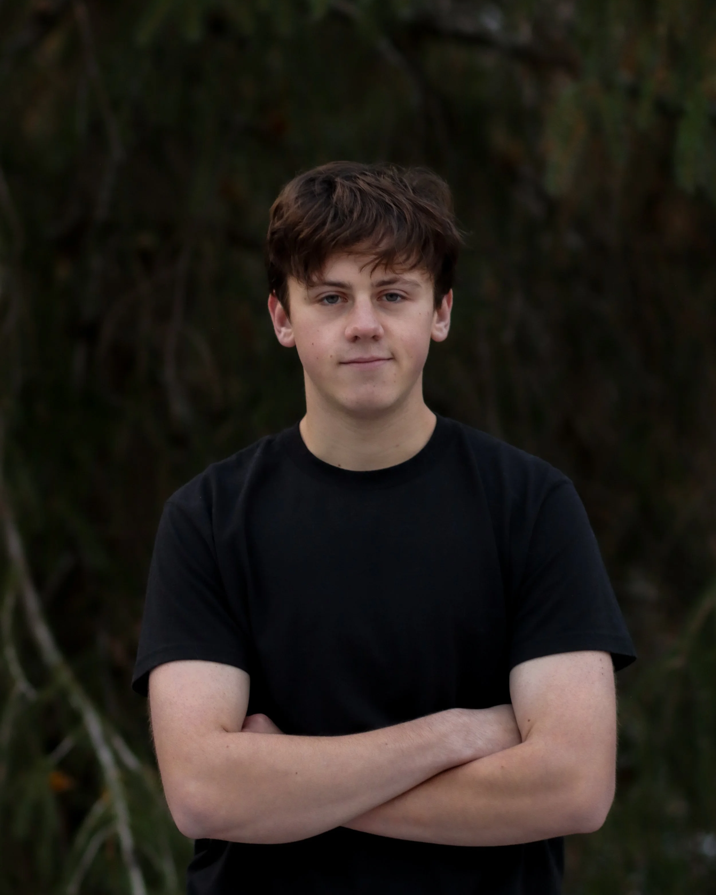 A teenage boy with brown curly hair wearing a black T-shirt standing outdoors with arms crossed in front of a hazy, dark green natural background.