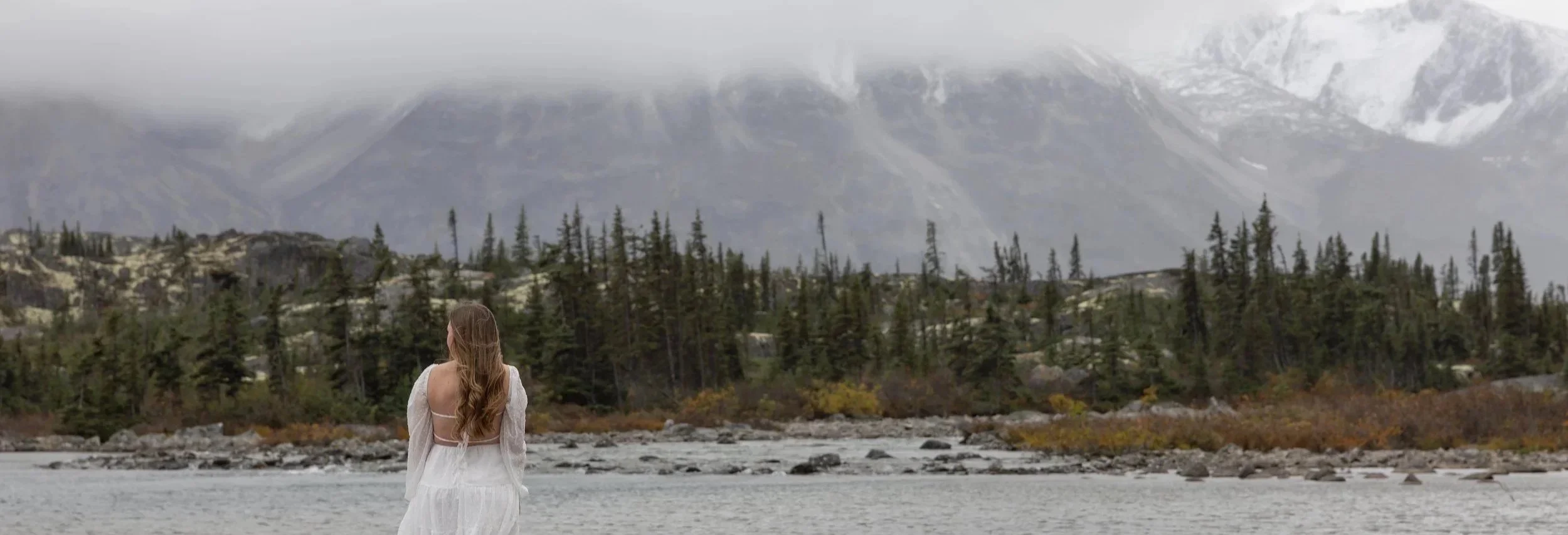 A woman with long hair in a white dress standing by a river, facing away, with trees, rocky terrain, and snow-capped mountains in the background.