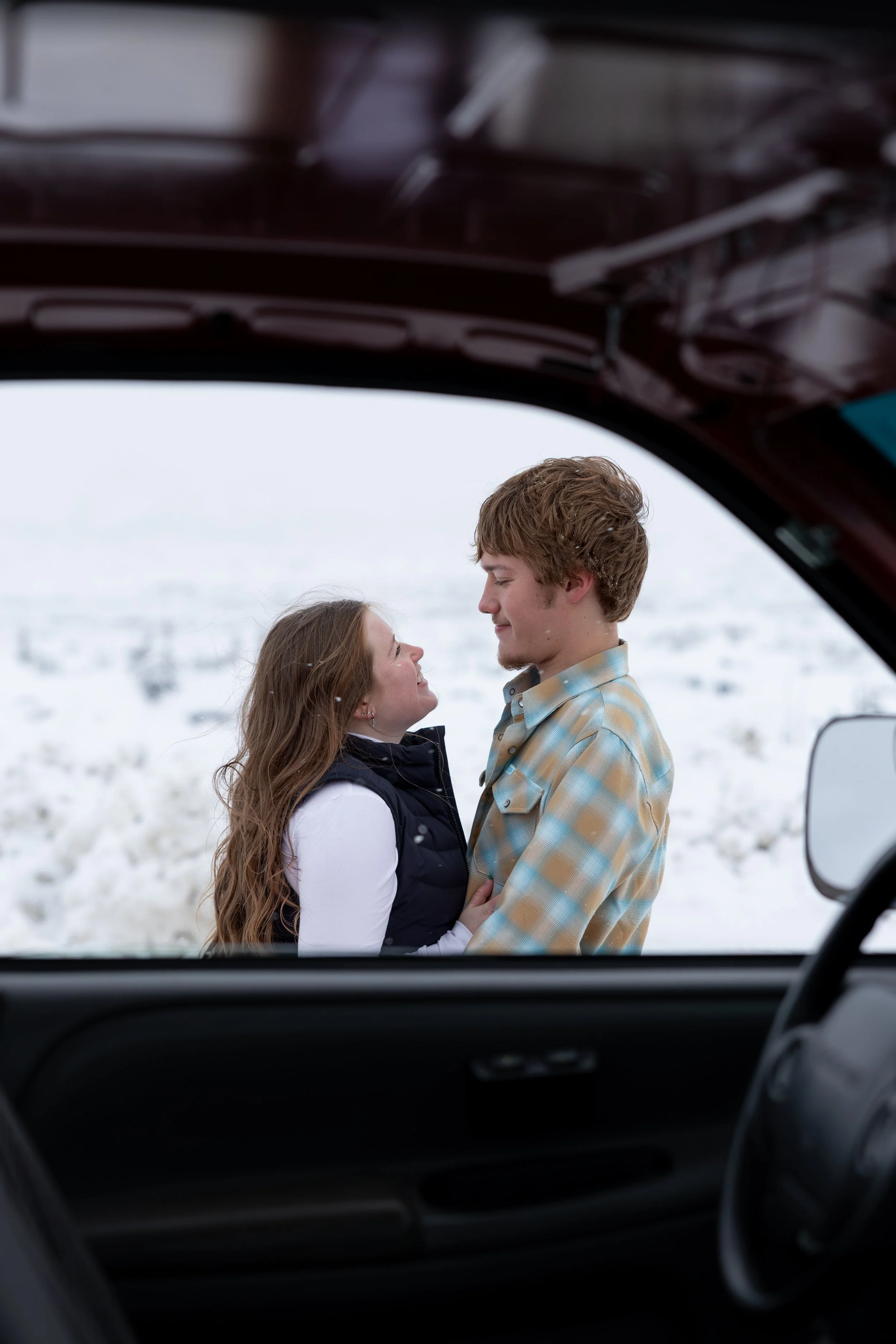 Young couple gazing at each other in a snowy landscape, viewed from inside a vehicle.