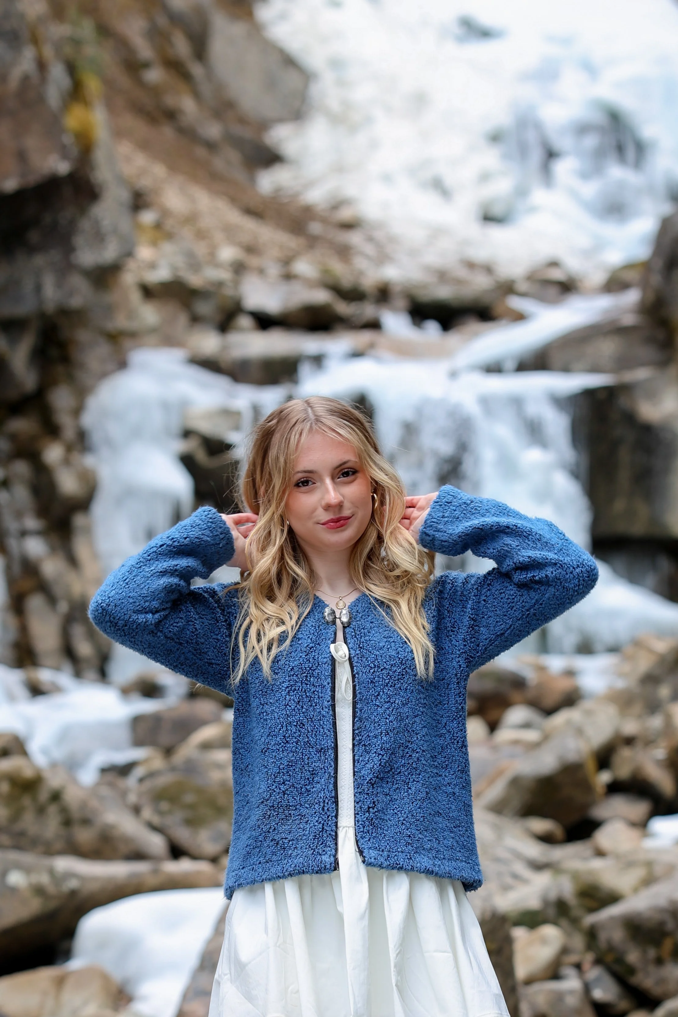 A young woman with blonde hair wearing a blue jacket and white dress standing in front of a waterfall with snow and rocks.