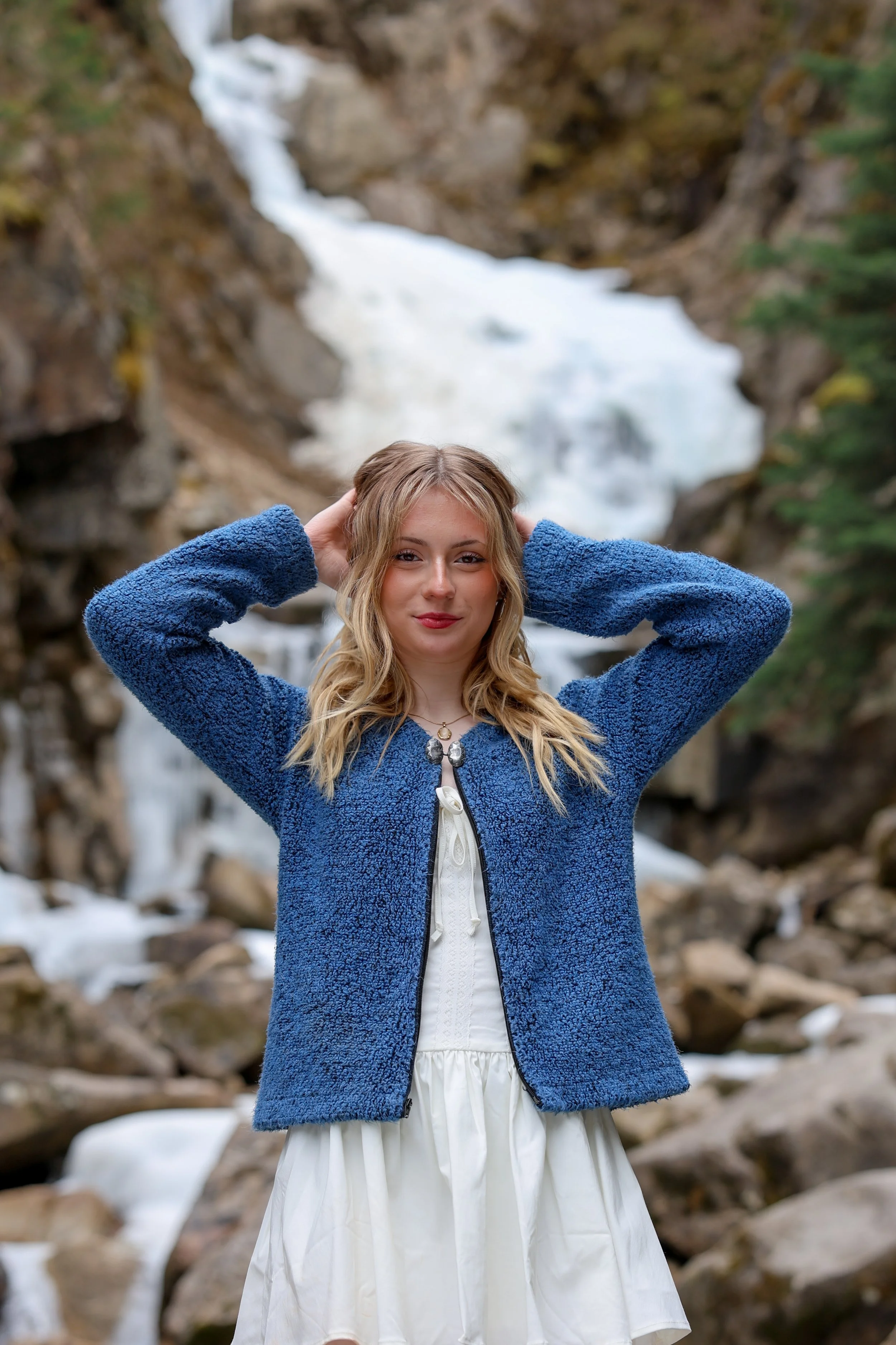 A young woman in a white dress and blue fleece jacket stands outdoors in front of a flowing waterfall surrounded by rocks and trees, posing with her hands behind her head.