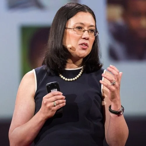 Woman with shoulder-length dark hair, glasses, and a pearl necklace speaking at a conference, holding a remote control in her right hand.