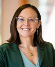 Portrait of a woman with brown hair, wearing clear glasses, a green blazer, and a blue top, smiling indoors.