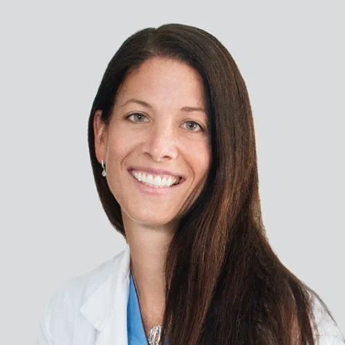 Professional woman with long dark hair, smiling, wearing a white lab coat and a light blue shirt, against a light gray background.