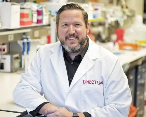 A smiling man in a lab coat standing in a laboratory with shelves of supplies in the background.