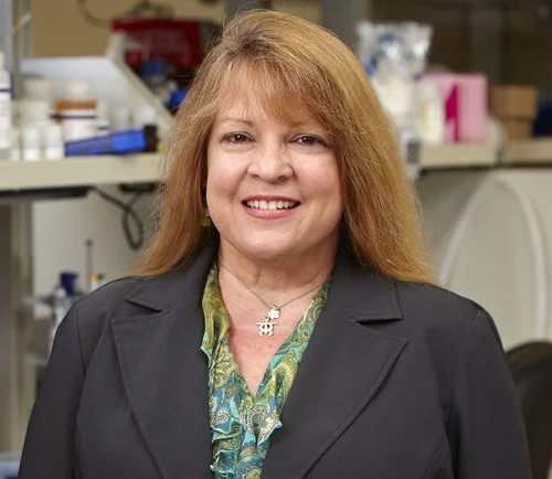Portrait of a woman with red hair smiling, wearing a dark blazer and colorful blouse, in a laboratory setting.