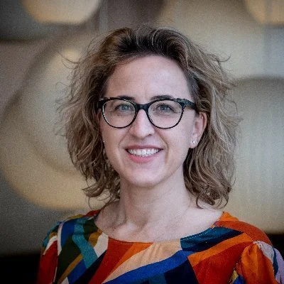 A woman with curly hair and glasses smiling for the camera, wearing a colorful patterned top.