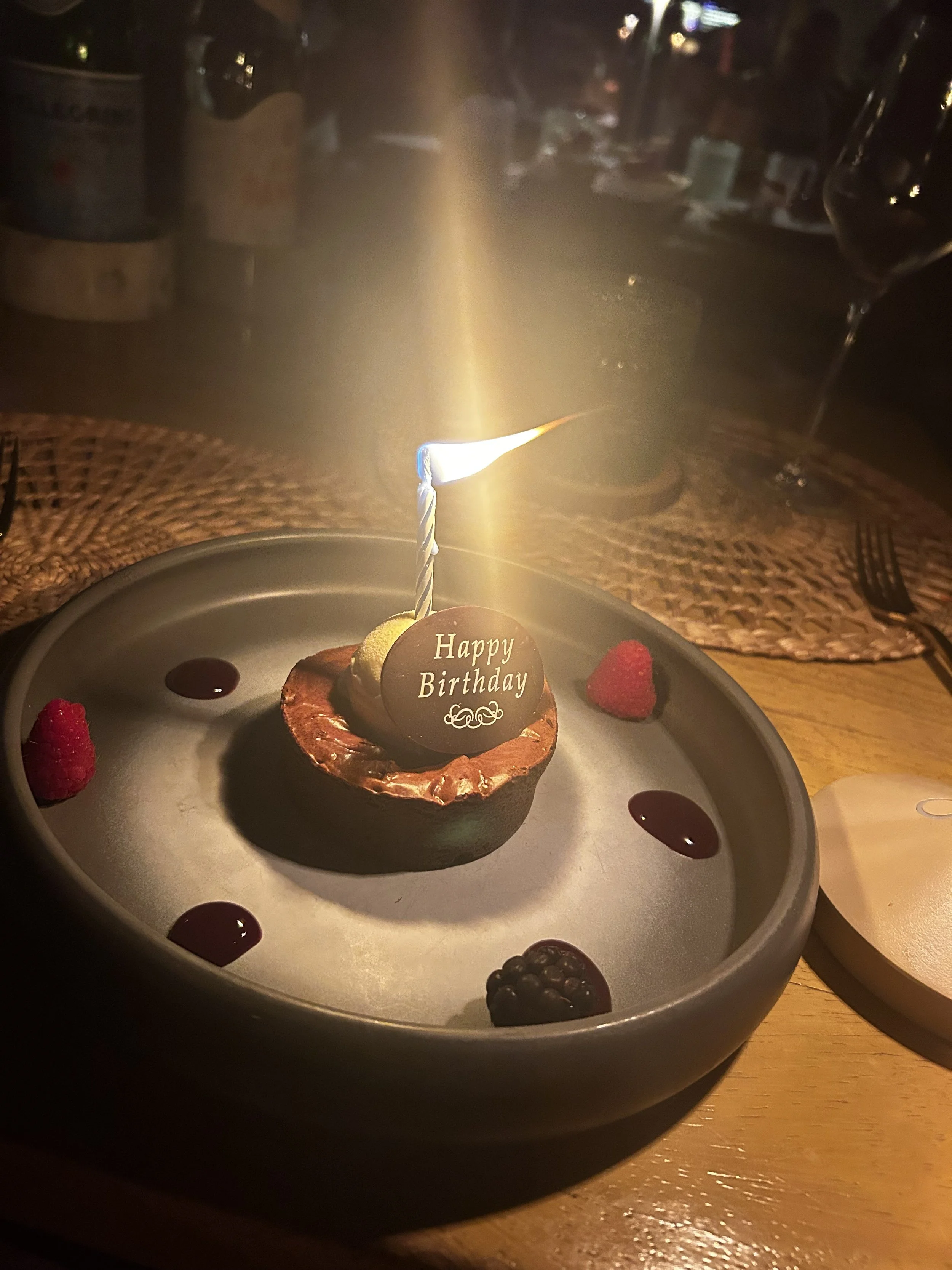 Small chocolate birthday dessert on a gray plate with a single lit candle, ‘Happy Birthday’ chocolate plaque, and scattered berries in a dimly lit restaurant.