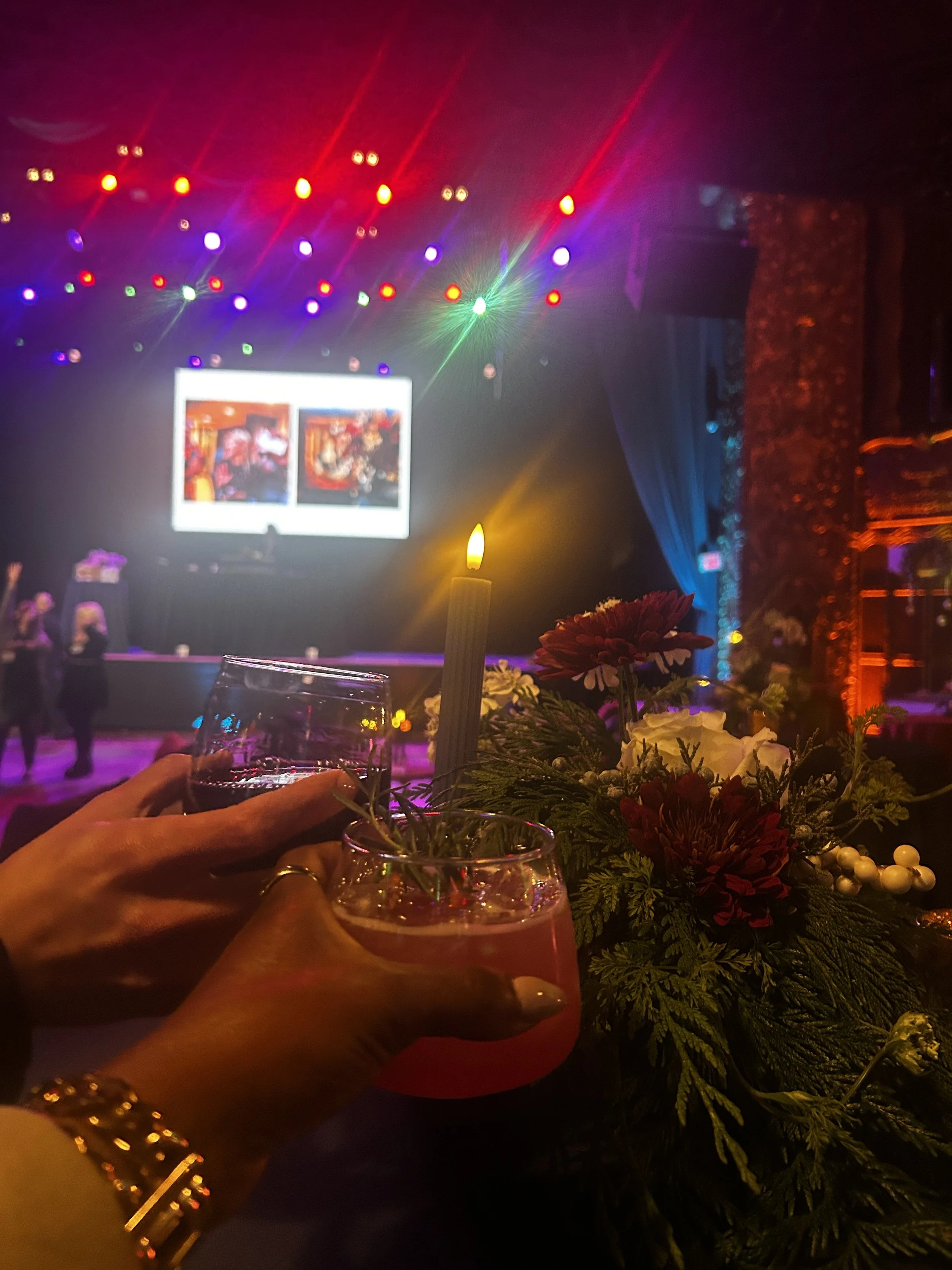 Hands clinking glasses in front of a stage lit with colorful lights at an evening event.