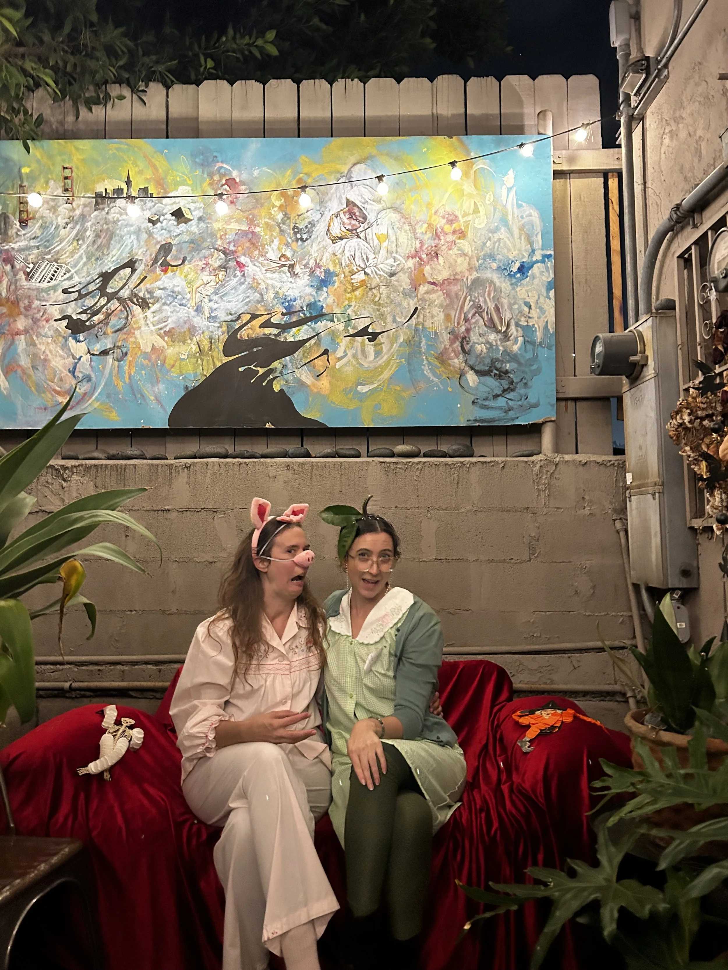Two people in playful costume makeup sitting on a red velvet couch beneath a vibrant abstract painting and string lights, capturing the whimsical, art-house energy of Shy Rockstar Studios.