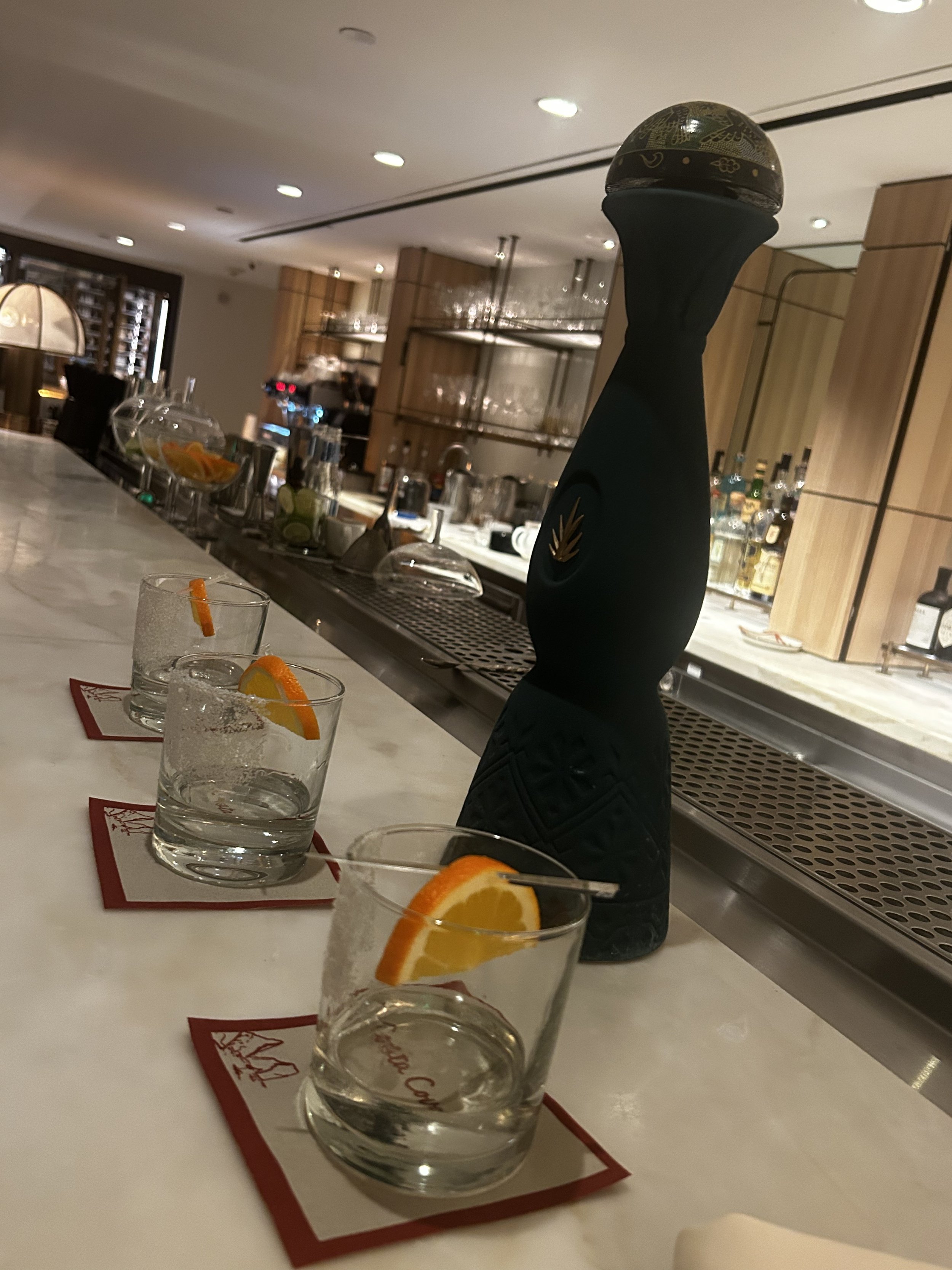 Tequila bottle and glasses lined up on a marble bar counter in a softly lit upscale venue.