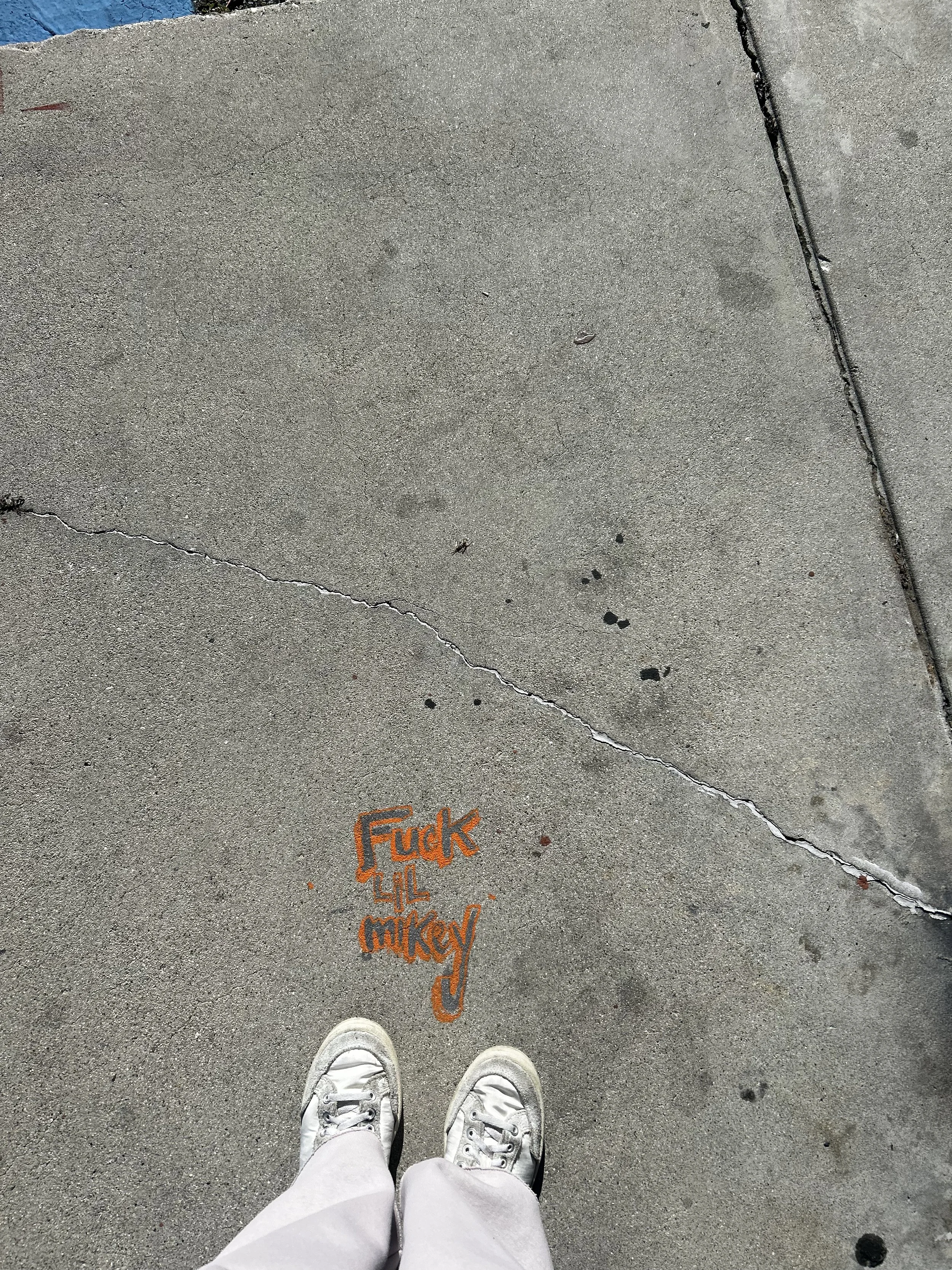 Looking down at worn sneakers on a city sidewalk with handwritten chalk text, capturing an unfiltered urban moment.
