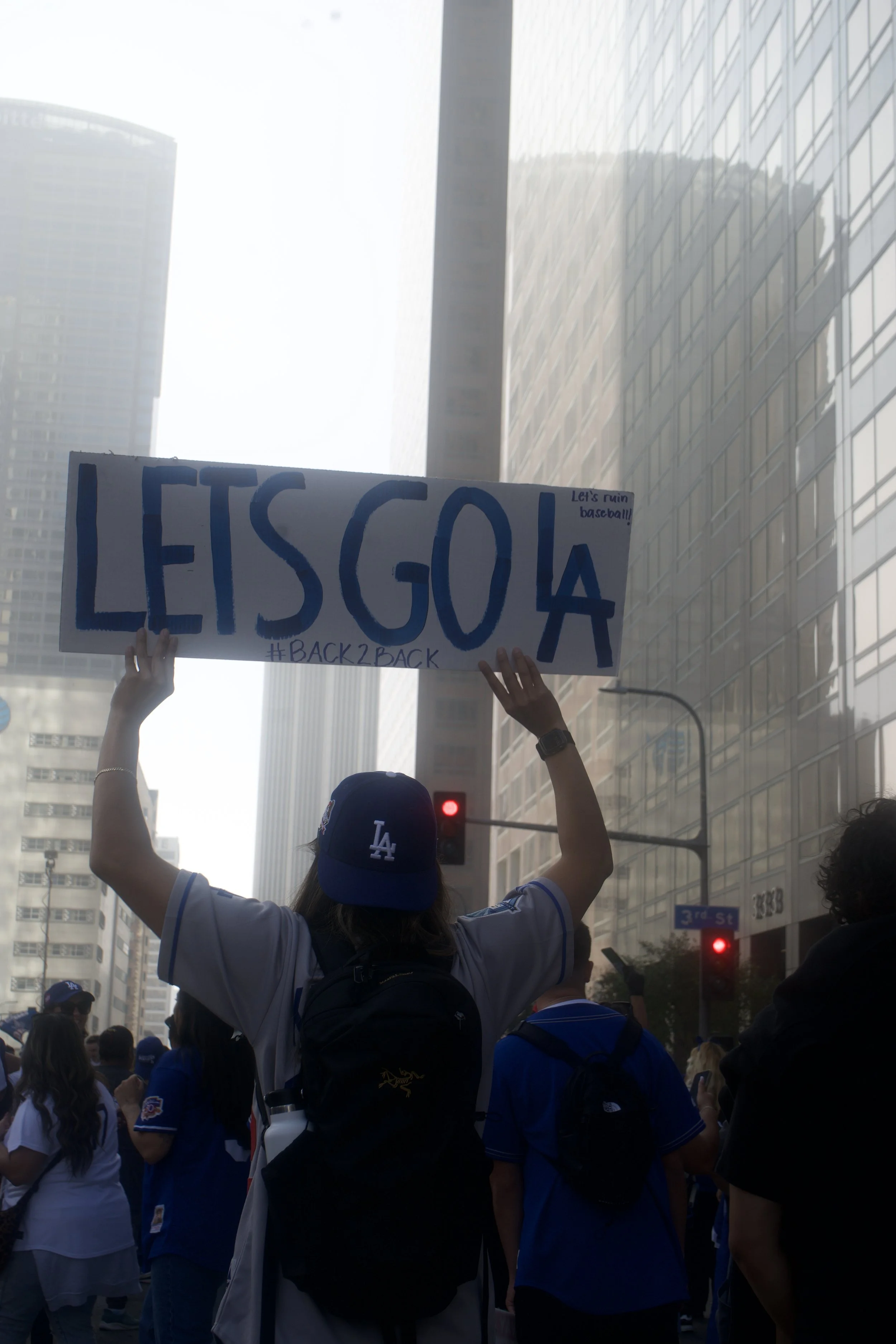 Person in a Dodgers jersey holding a “LET’S GO LA” sign above their head in the middle of a crowded downtown street, capturing the celebratory, city-energy mood of Shy Rockstar Studios.
